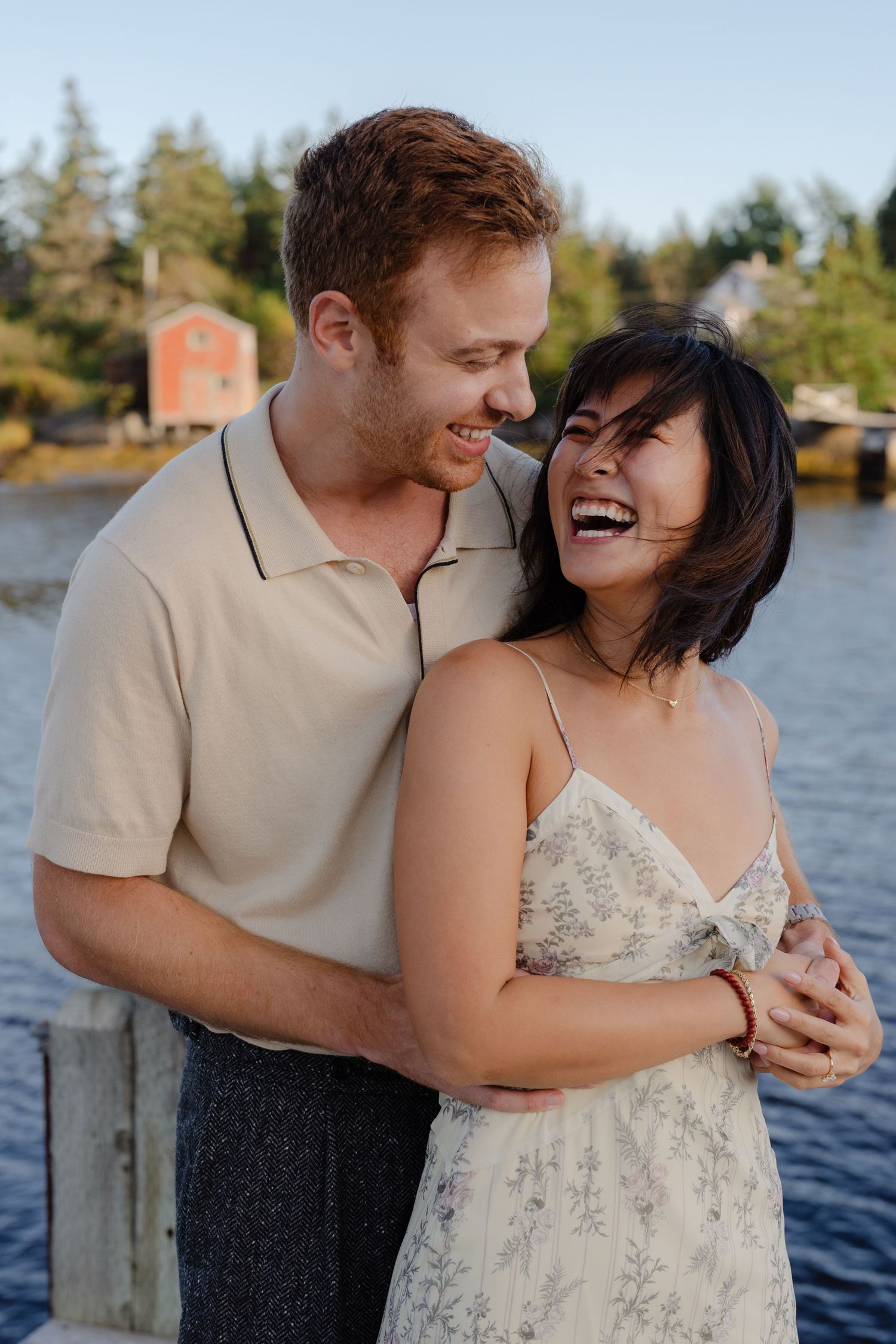 Happy couple embracing and laughing together on a waterfront dock with trees and a red boathouse behind them.