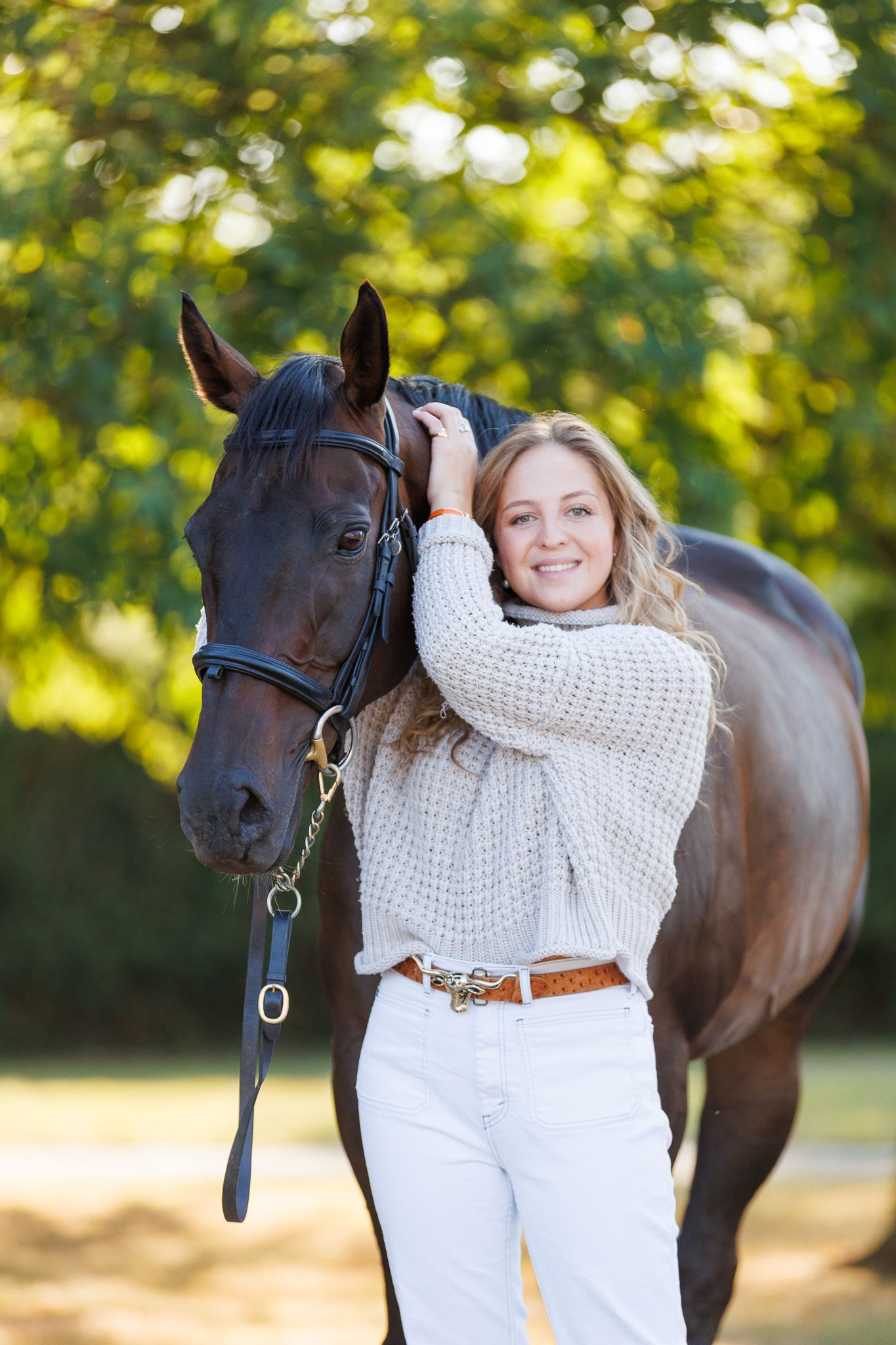 Young woman in navy blouse sits on bench between two bay horses with white markings, posed against brick wall.