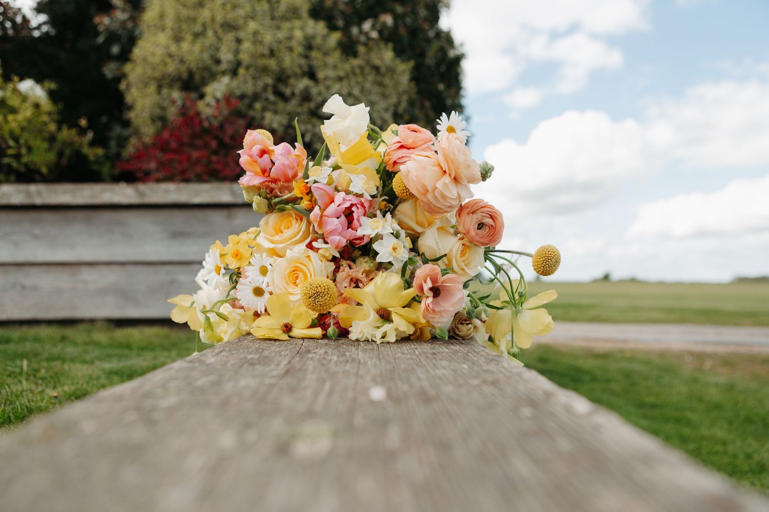 Pastel bridal bouquet of peach, pink, white and yellow flowers resting on a weathered wooden beam overlooking a green field and cloudy sky.