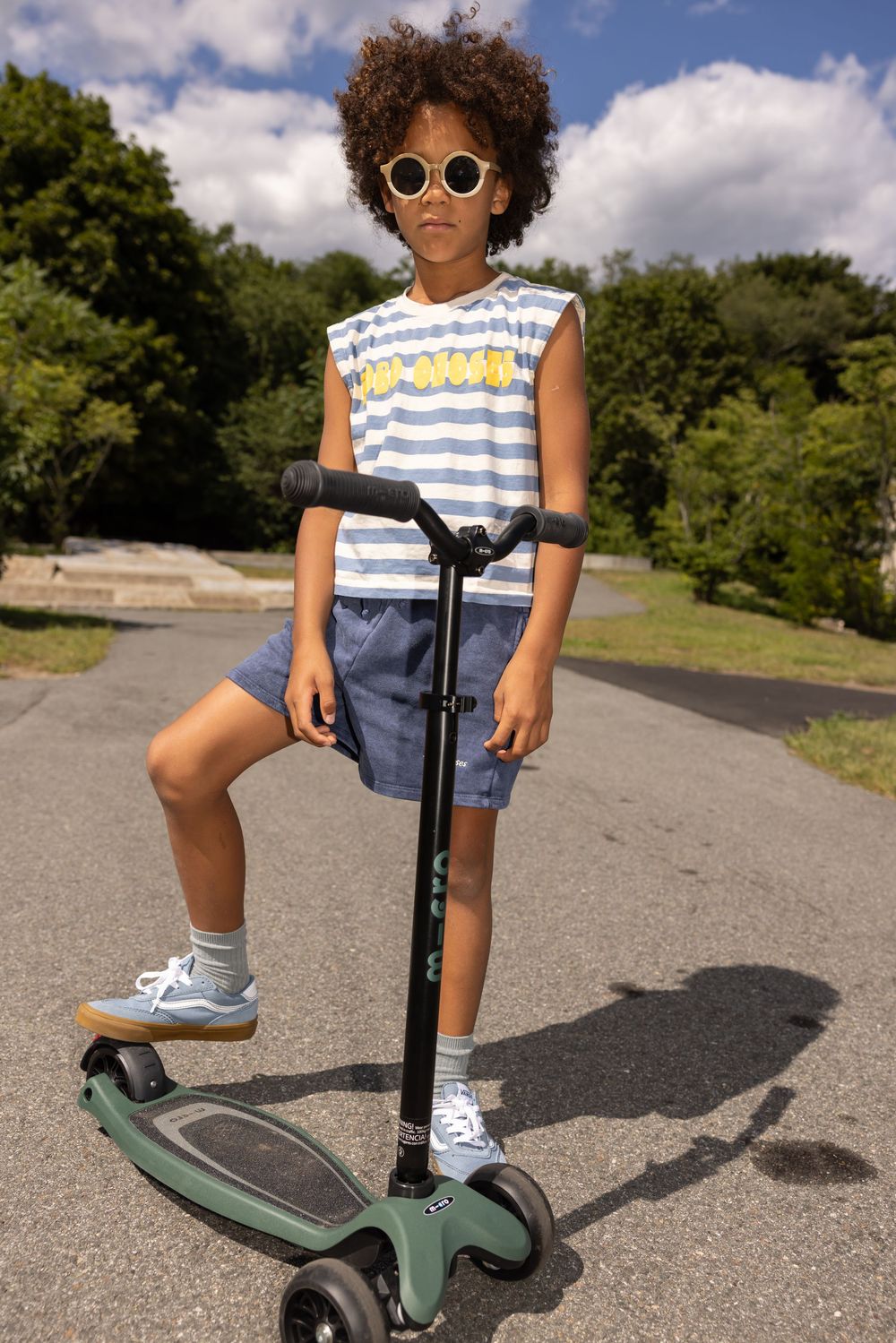 Child with curly hair wearing sunglasses, striped tank top, and shorts, riding a green kick scooter outdoors.