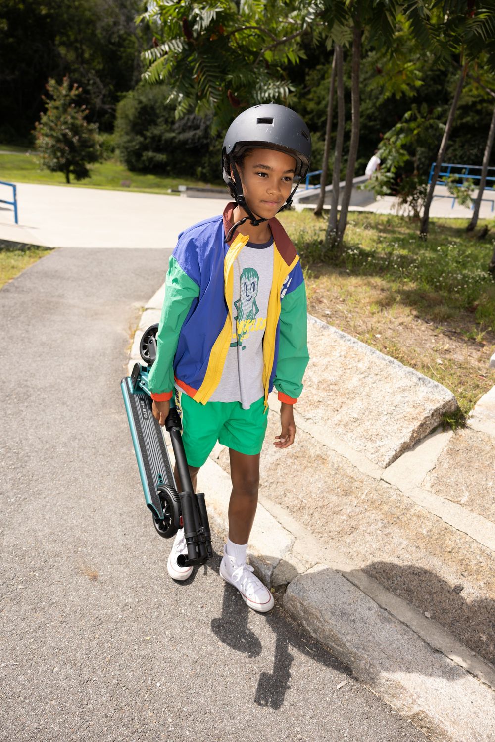 Young boy in colorful jacket, helmet, and green shorts carrying a scooter in a sunny park.