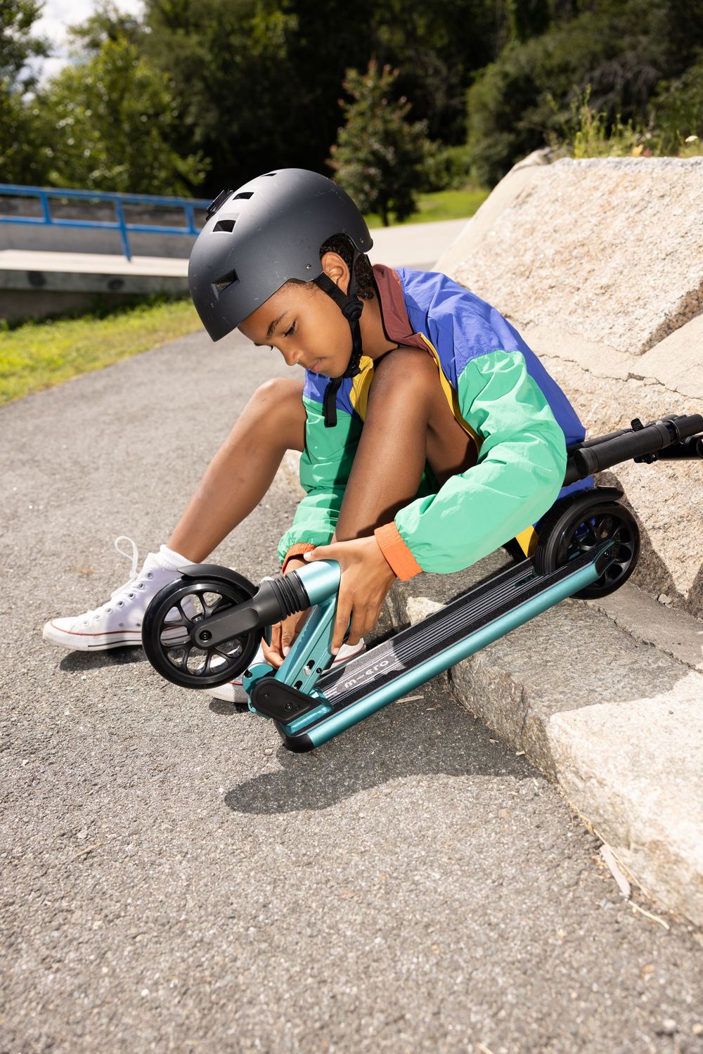 Young child wearing a helmet adjusts their teal kick scooter on a park path, dressed in colorful athletic wear.