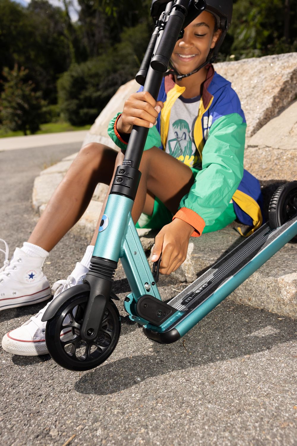 Smiling teen in colorful jacket and helmet sits on teal kick scooter on pavement in a park.