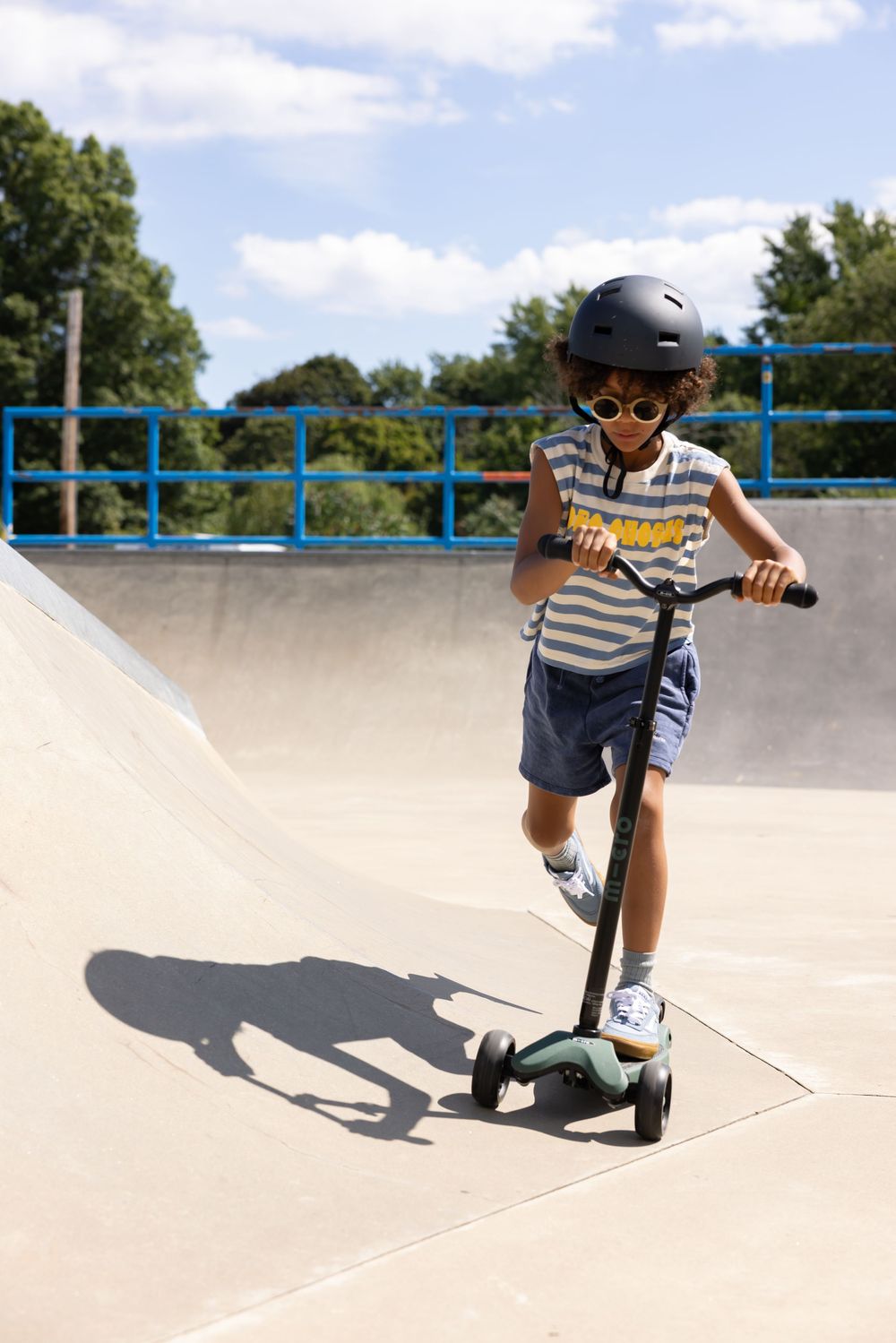 A young child wearing a helmet and sunglasses rides a kick scooter at a sunny outdoor skate park.