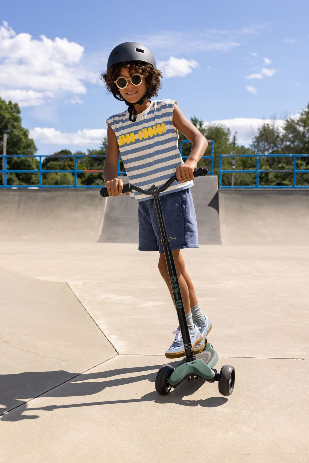 A smiling child wearing a helmet and sunglasses rides a green kick scooter at a sunny outdoor skate park.