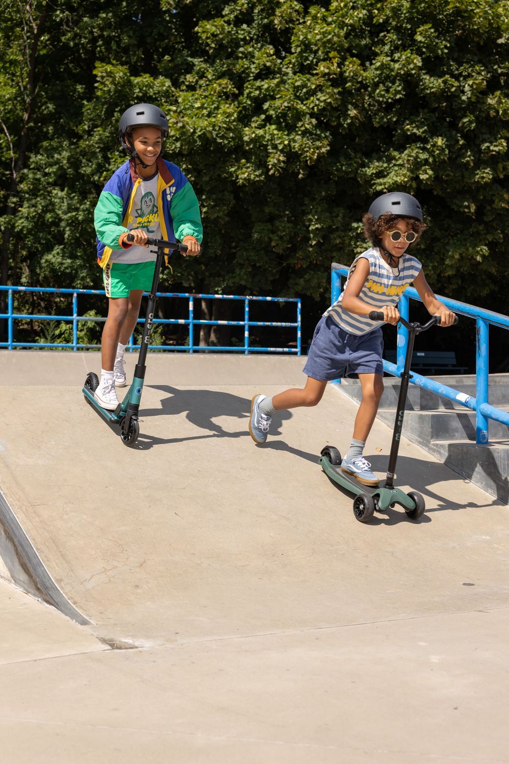 Two kids wearing helmets ride scooters down a ramp at a sunny outdoor skate park.