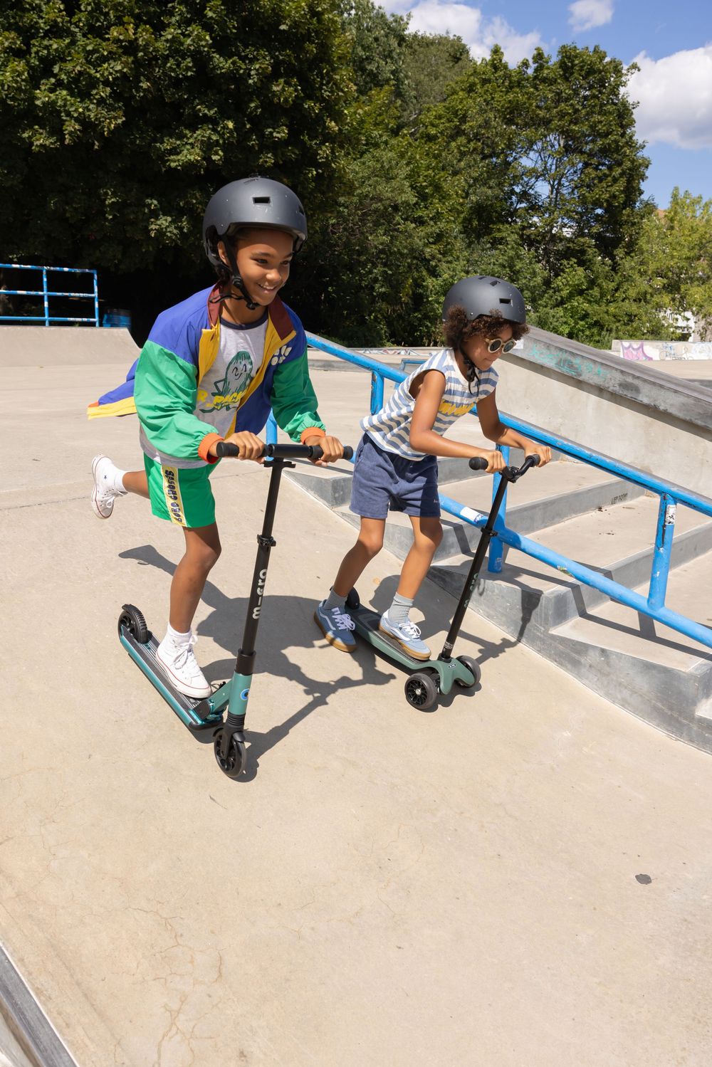 Two kids wearing helmets ride scooters down a ramp at a sunny skate park.
