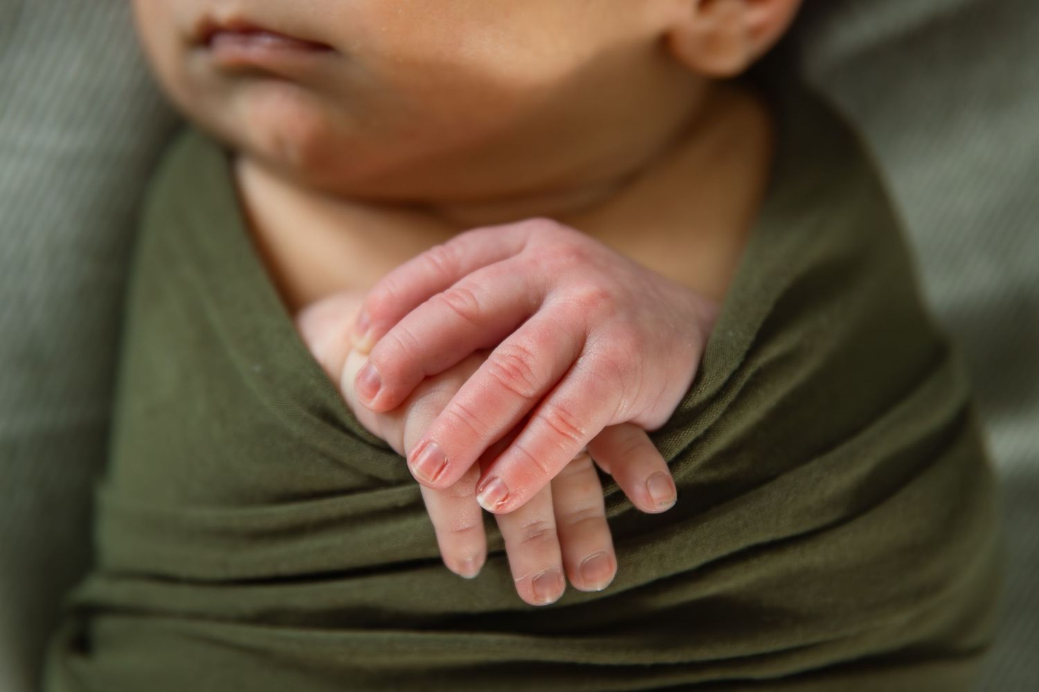 Close-up of a newborn baby's tiny hand resting on an olive green swaddle wrap.