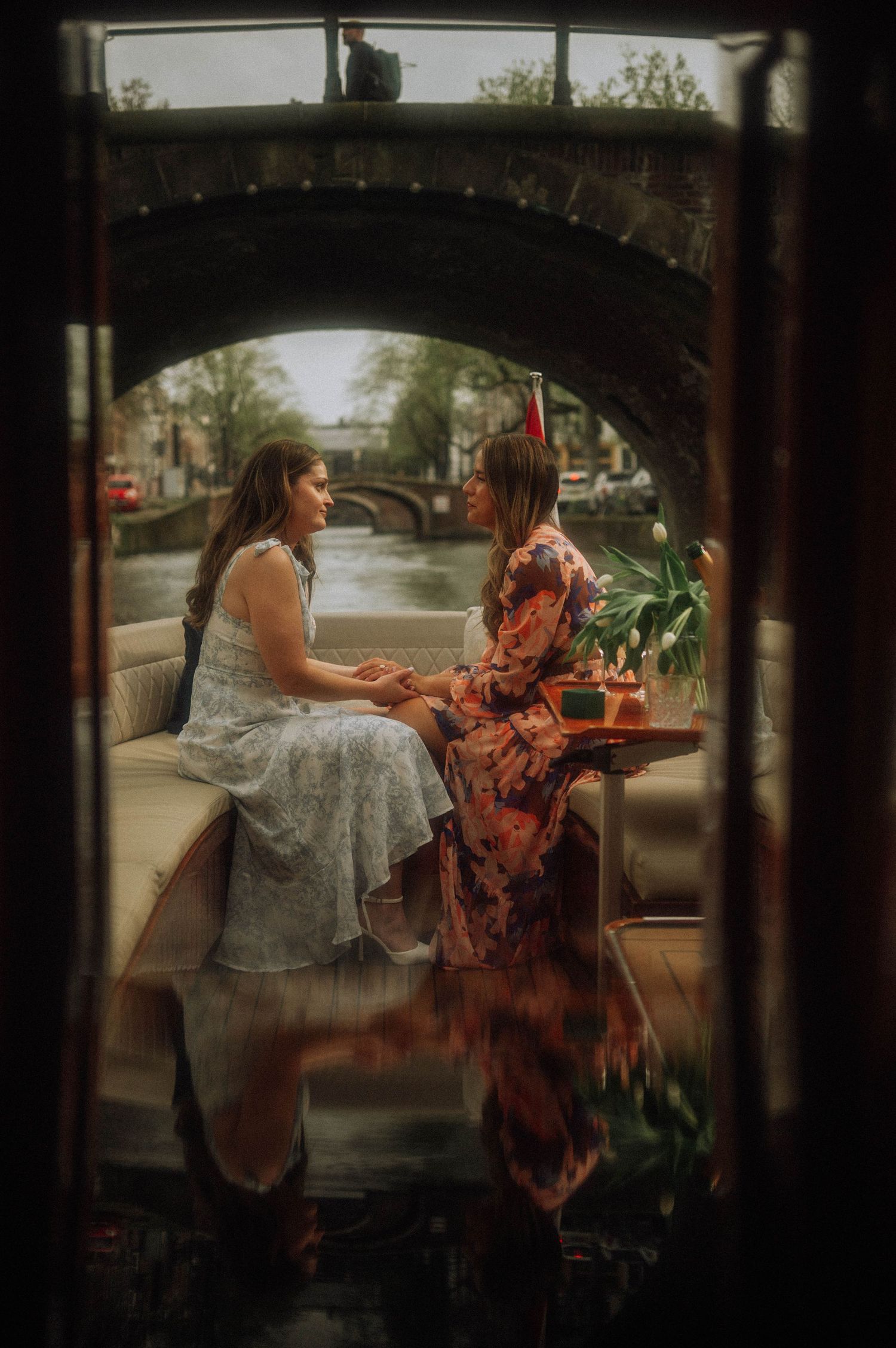 Two women in floral dresses sit holding hands on a canal boat, with a stone bridge and flowers on the table behind them. Amsterdam