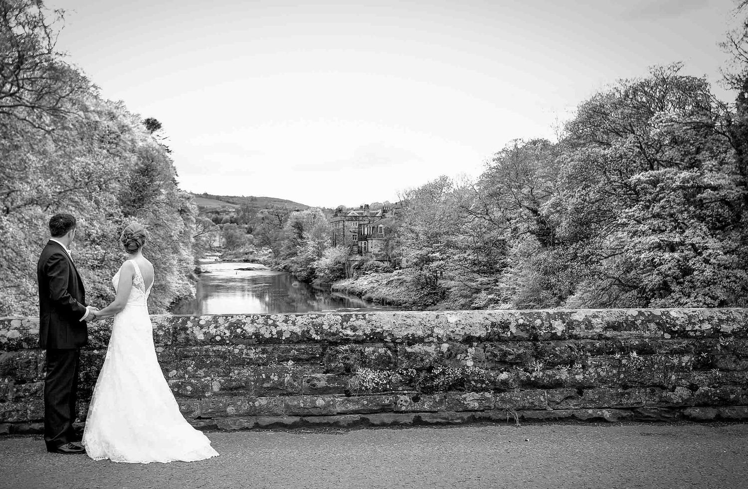 Black and white wedding portrait of the bride and groom on a stone bridge overlooking a river and wooded hills.