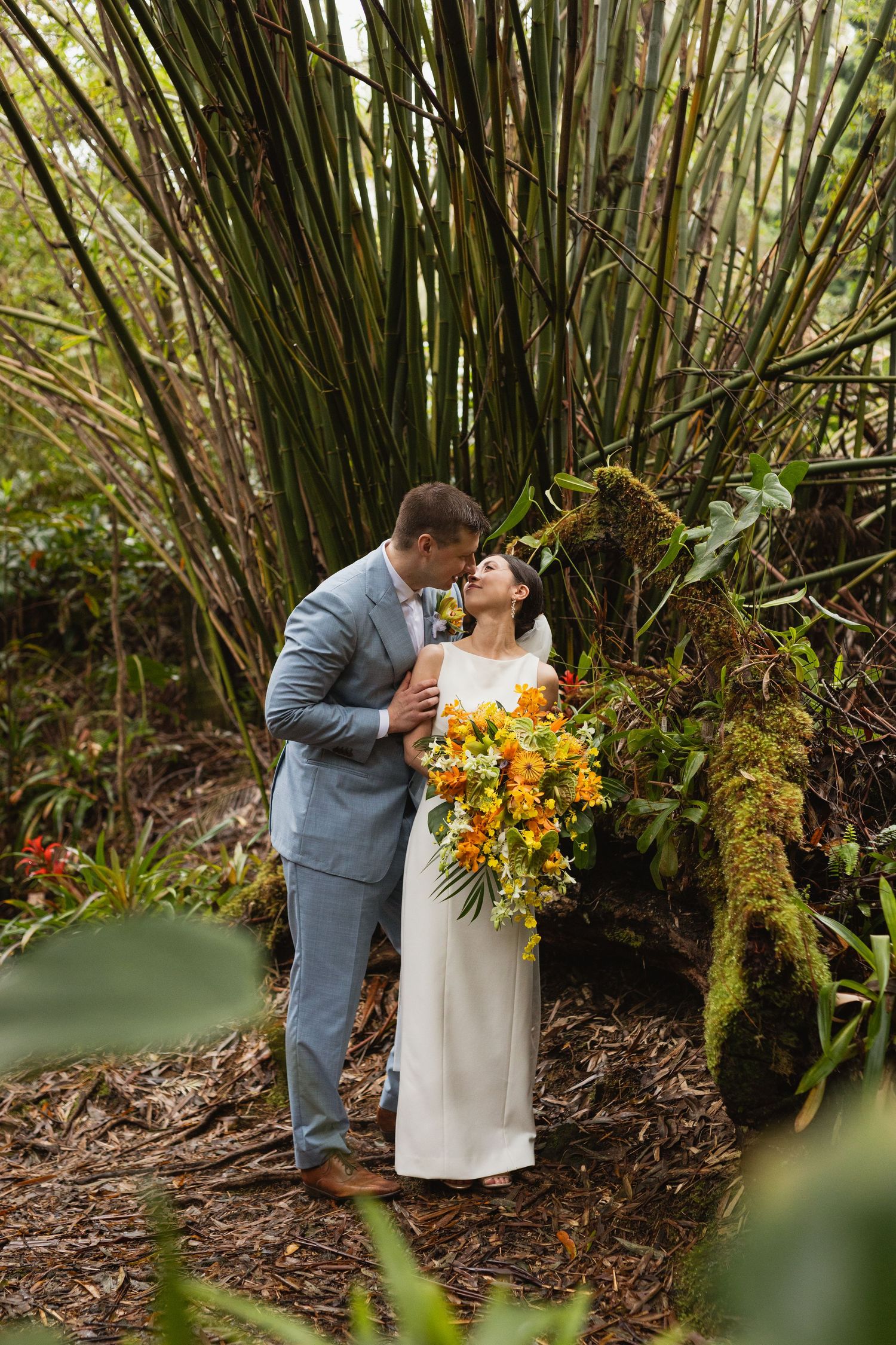 Bride and groom embrace among towering bamboo and moss-covered logs, she holding a vibrant orange tropical bouquet in a lush forest wedding.