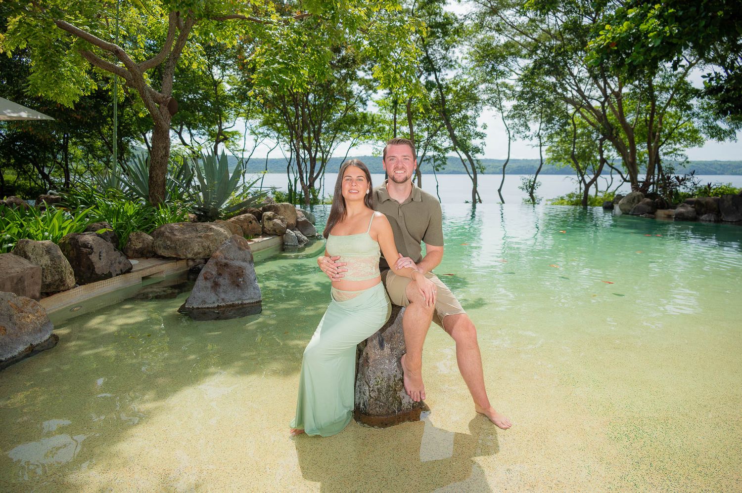 Smiling couple sits together on a rock in a clear shallow tropical pool surrounded by lush green trees, the woman in a flowing mint dress.