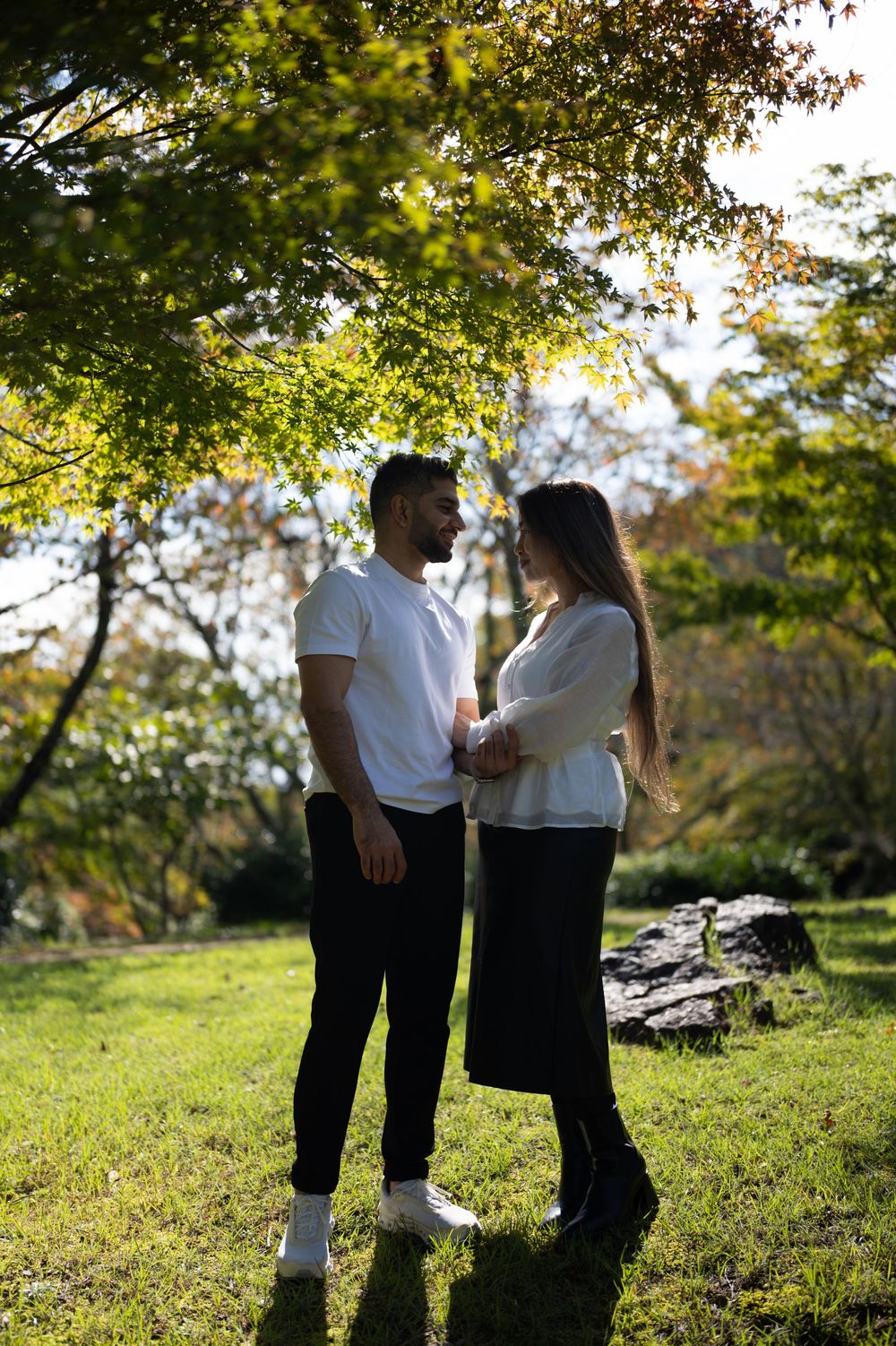 Couple standing face to face holding hands on a sunlit grassy lawn beneath green maple trees in a peaceful park.