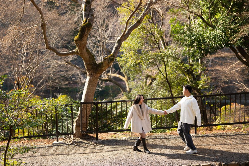 A couple in light coats holds hands and walks along a gravel path beside a fence, framed by autumn trees in a peaceful park.
