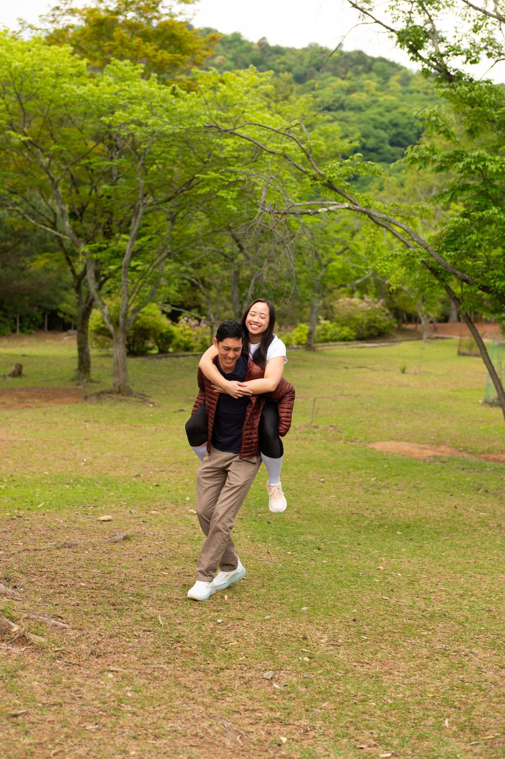 A smiling woman rides piggyback on a man walking across a sunny grassy park, with green trees and forested hills in the background.