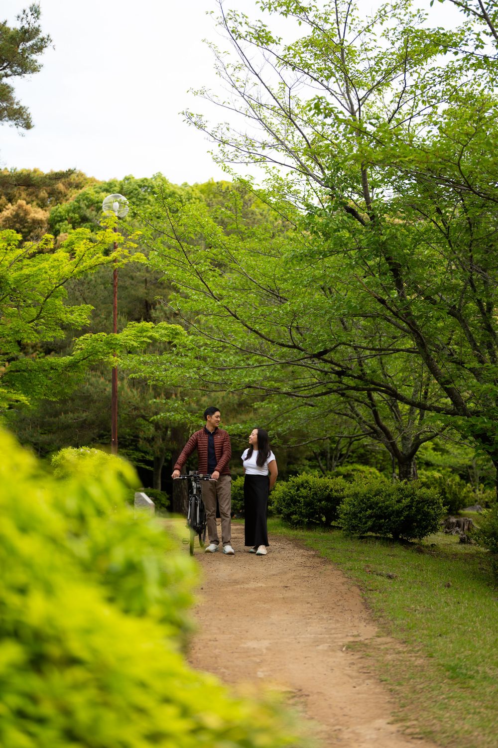 Couple walking along a dirt path through lush green trees and shrubs while the man pushes a bicycle on a sunny spring day.