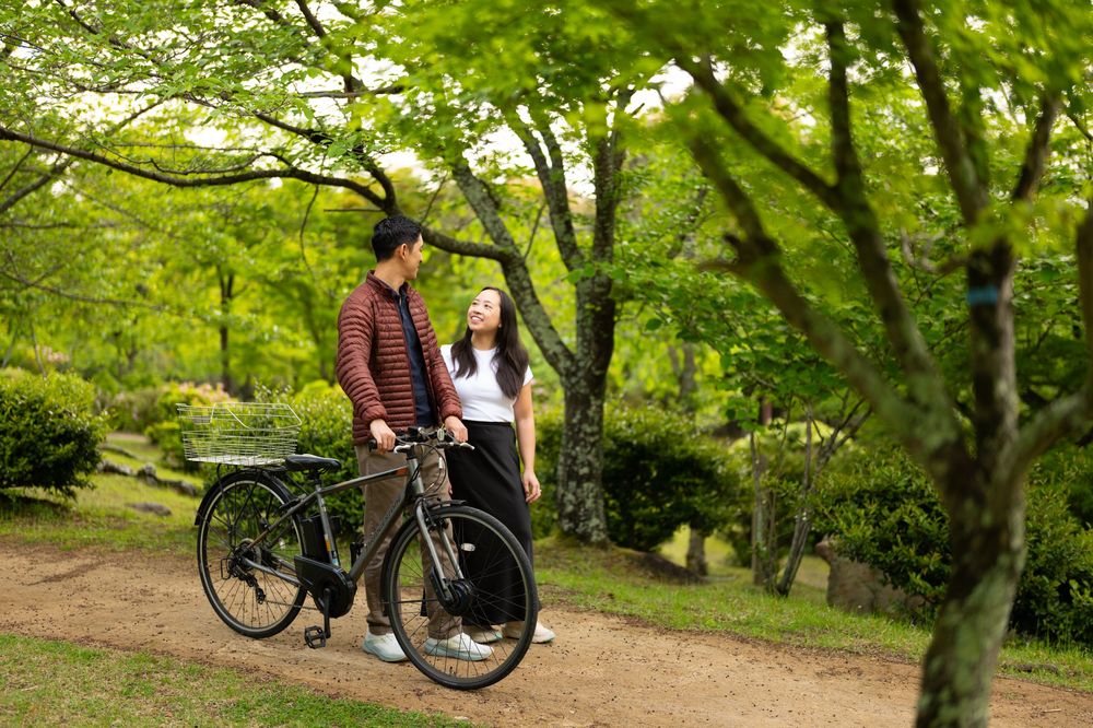 Couple standing beside a black bicycle on a dirt path in a lush green park surrounded by tall trees and shrubs.
