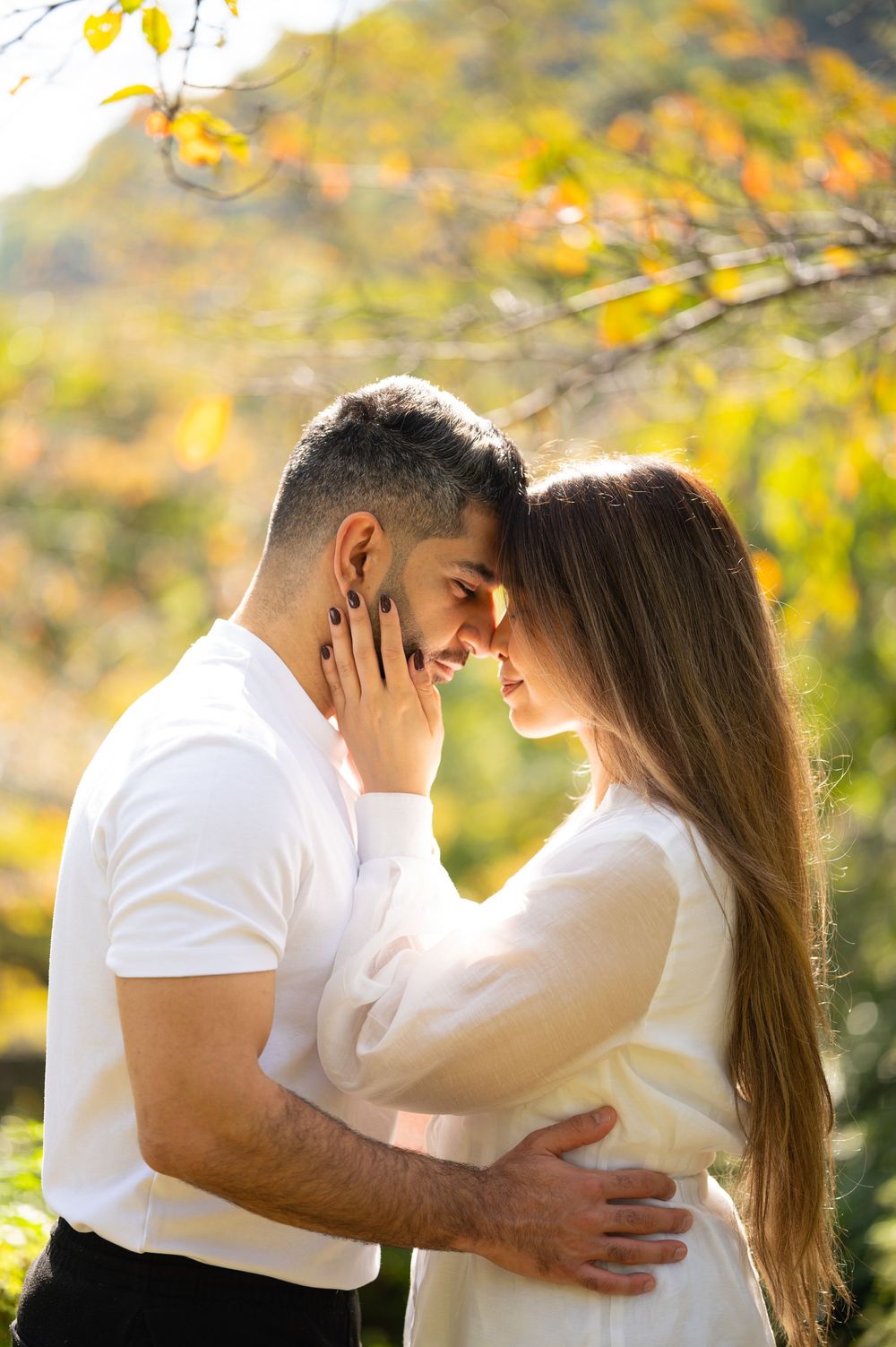 Couple in white outfits touching foreheads in a tender embrace outdoors, surrounded by blurred golden autumn foliage.
