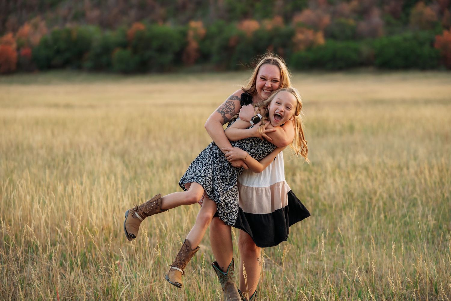 Two girls laughing and hugging in a golden field at sunset, wearing dresses and cowboy boots.