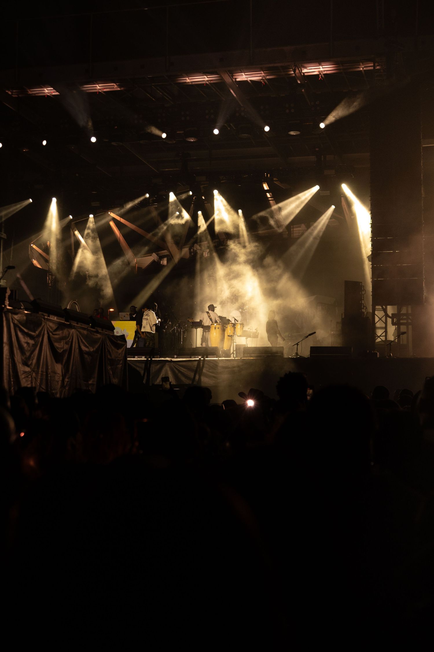 Live concert stage with dramatic yellow spotlights and smoke effects illuminating performers before a large crowd.