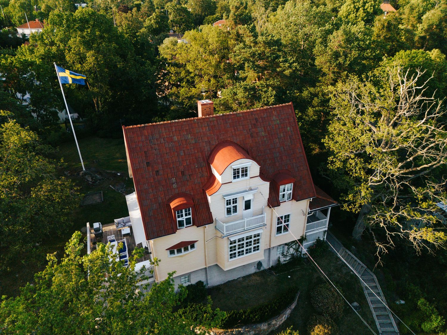 Aerial view of a charming cream villa with red tile roof surrounded by lush green trees on a sunny day.