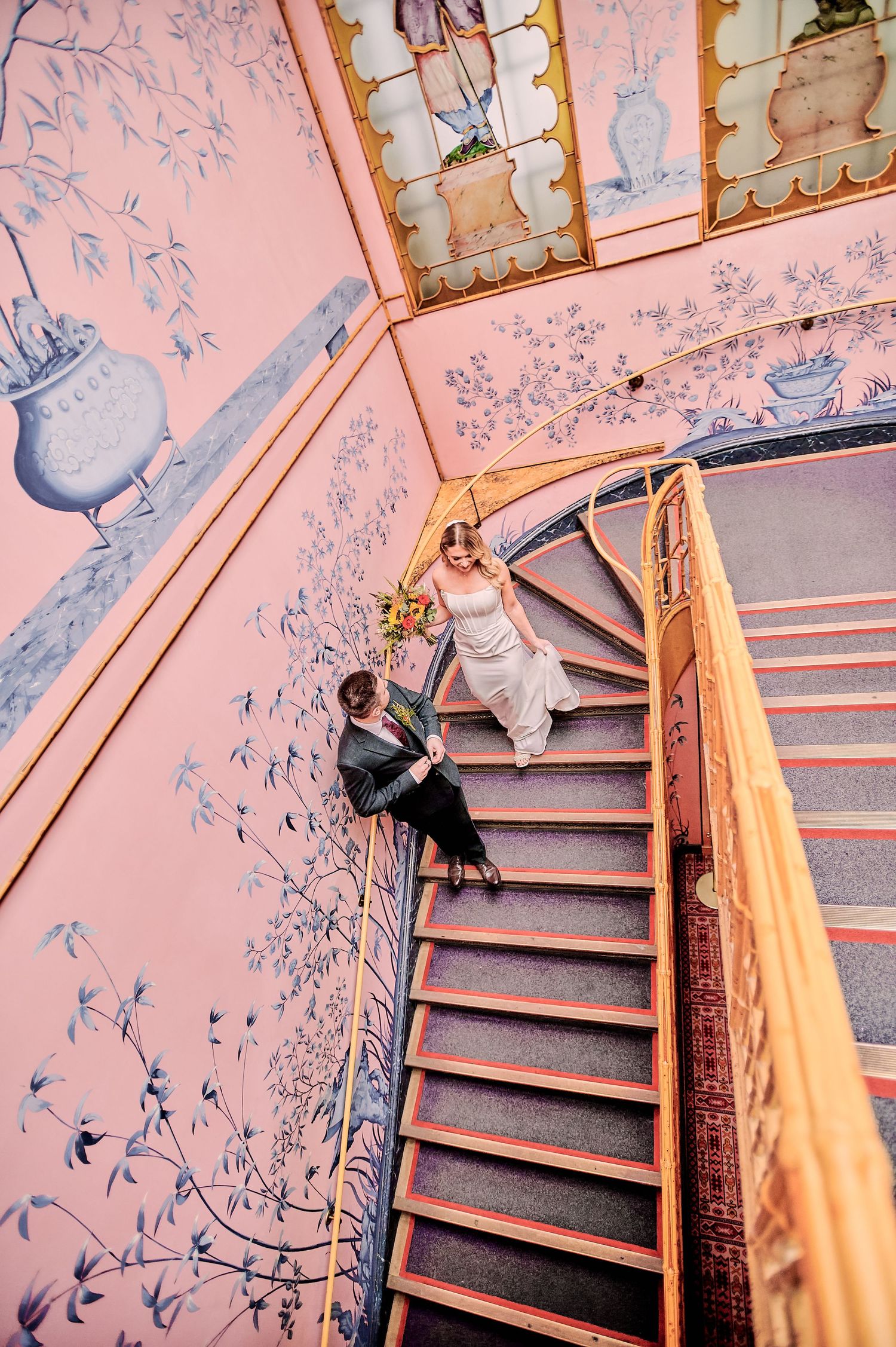 Bride and groom on ornate pink spiral staircase with blue floral murals, aerial view wedding photo.