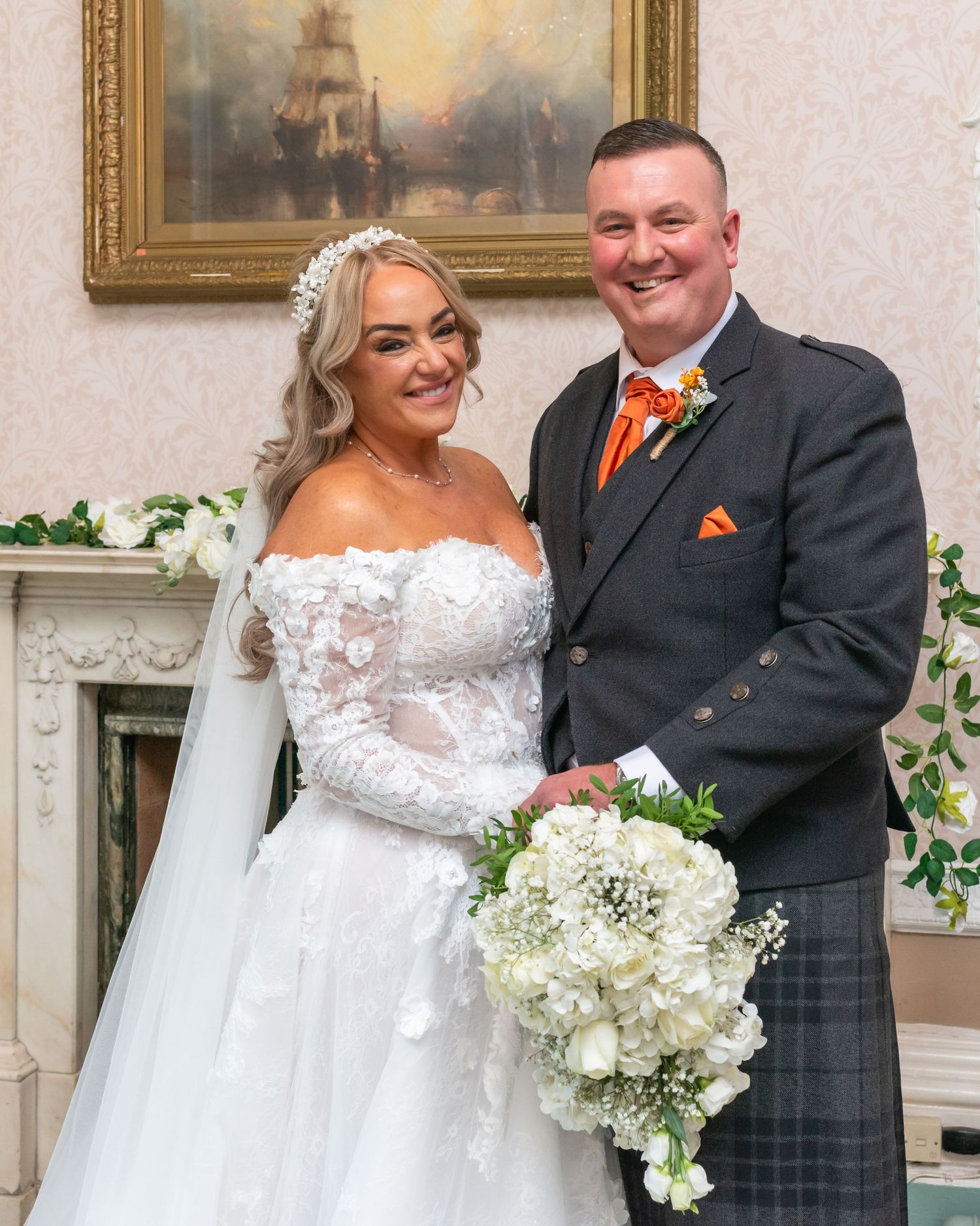 A smiling bride in a lace gown holds white flowers beside a groom in a grey tartan kilt with orange bow tie at Runcorn Town Hall