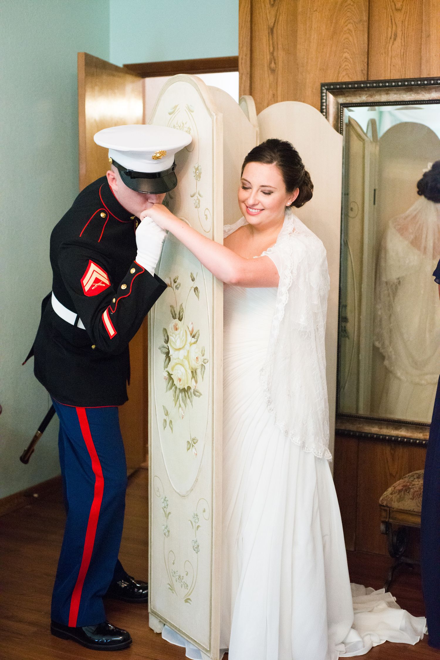 Marine in dress uniform kisses bride's hand beside decorative screen on their wedding day.