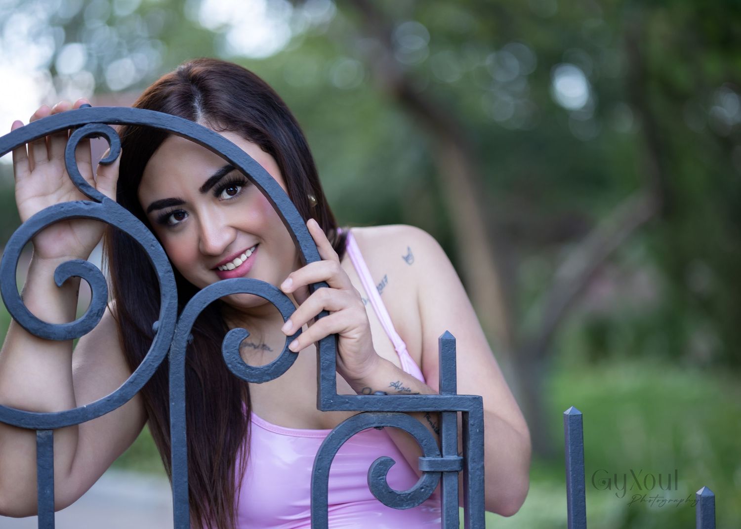Young woman with long brown hair smiling behind ornate iron gate in park, wearing pink top.