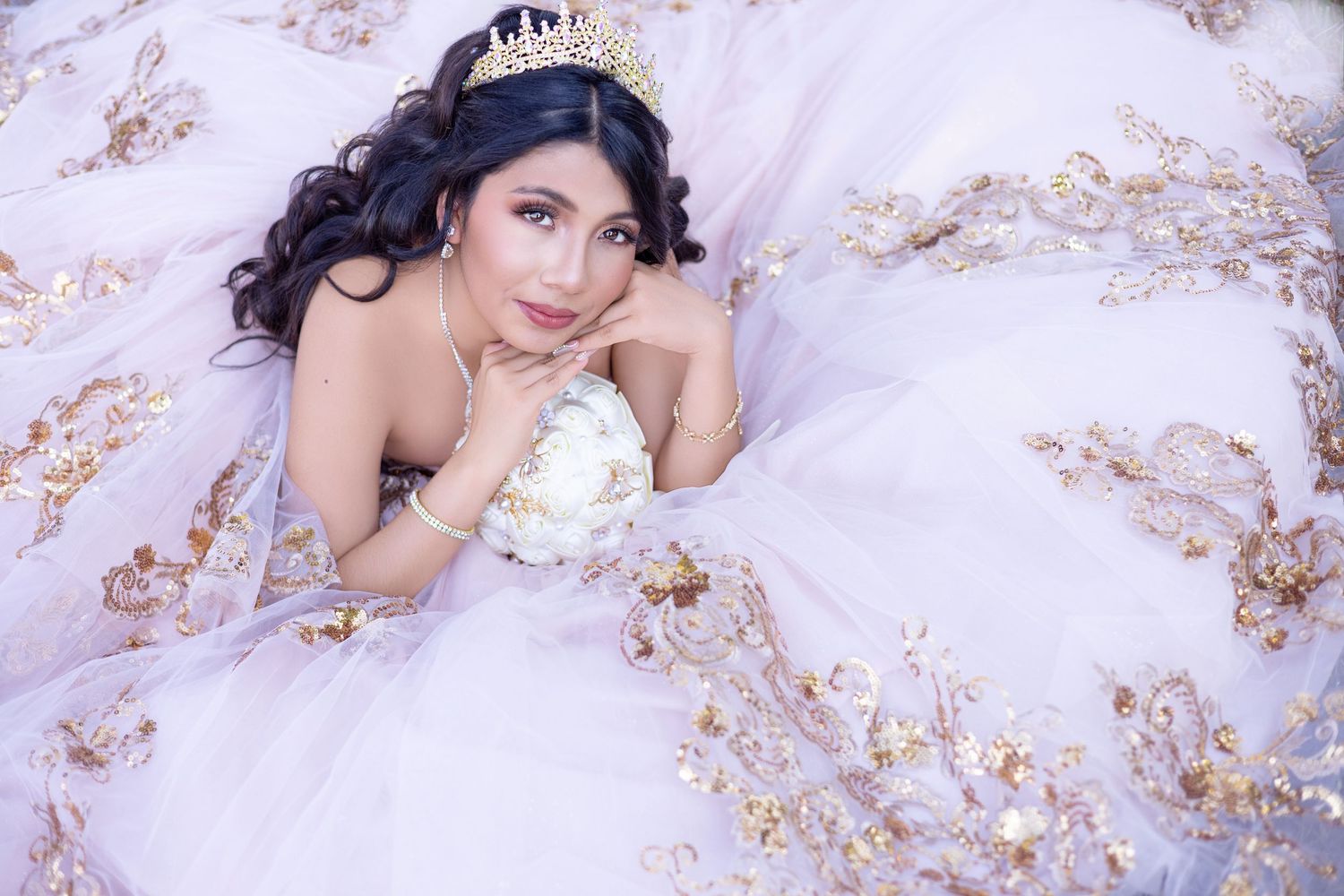 Young woman in ornate white and gold ballgown with tiara holding bouquet, lying elegantly for quinceañera portrait.