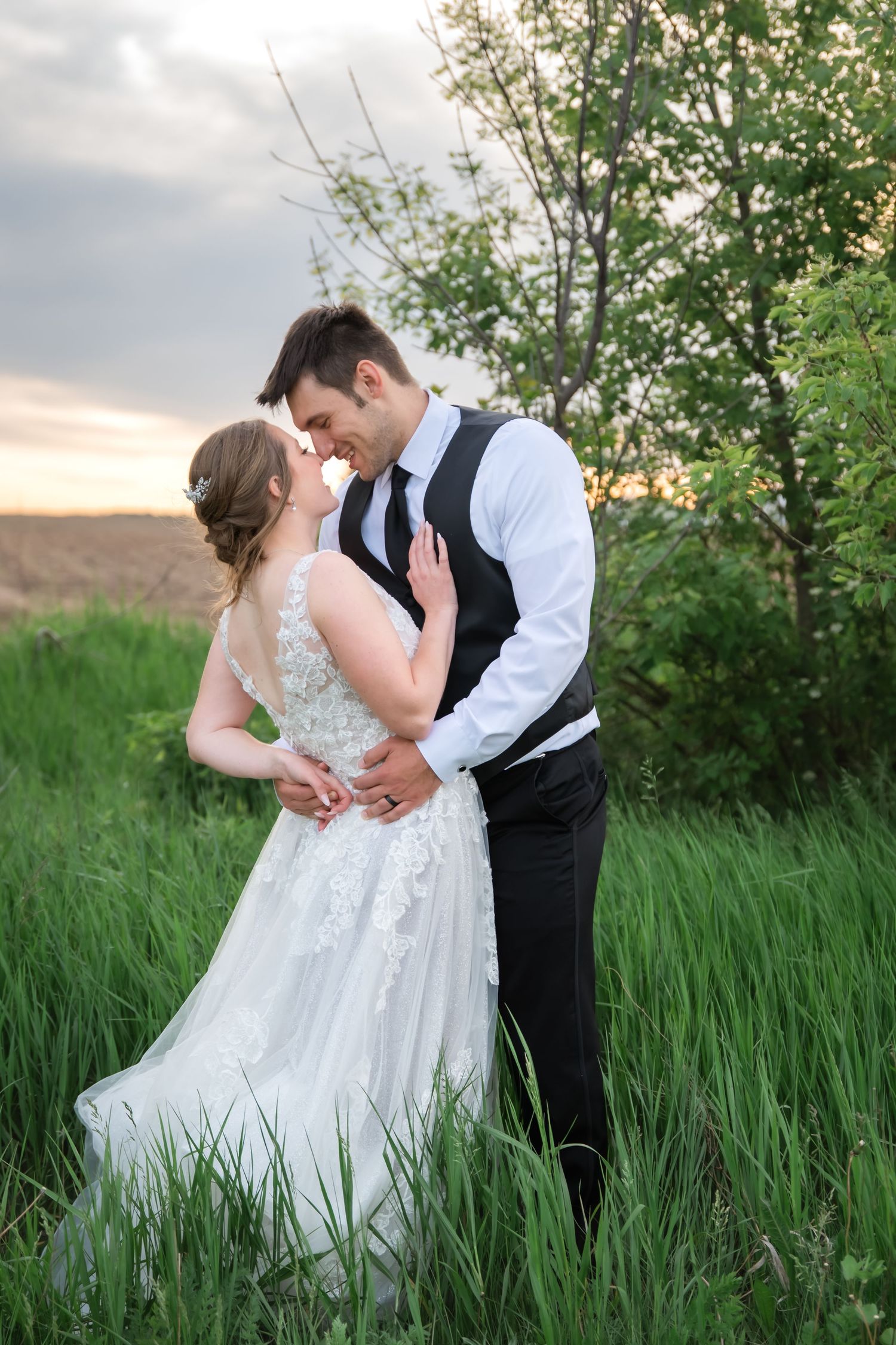 Bride in a lace wedding gown and groom in a black vest embrace in tall green grass at sunset during an outdoor wedding photo session.