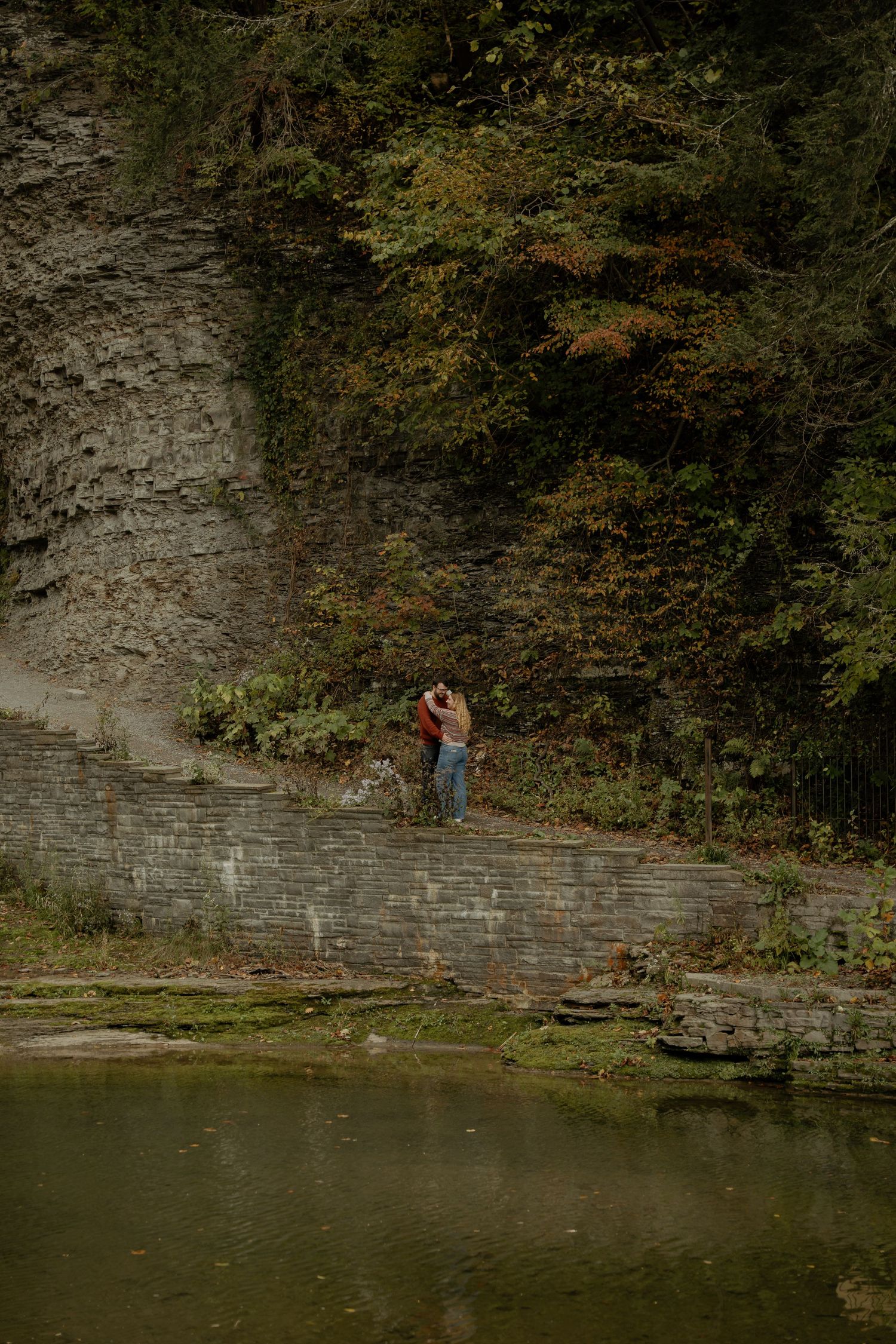 Couple embracing on a stone path beside a gorge wall and autumn foliage, with calm green water in the foreground.