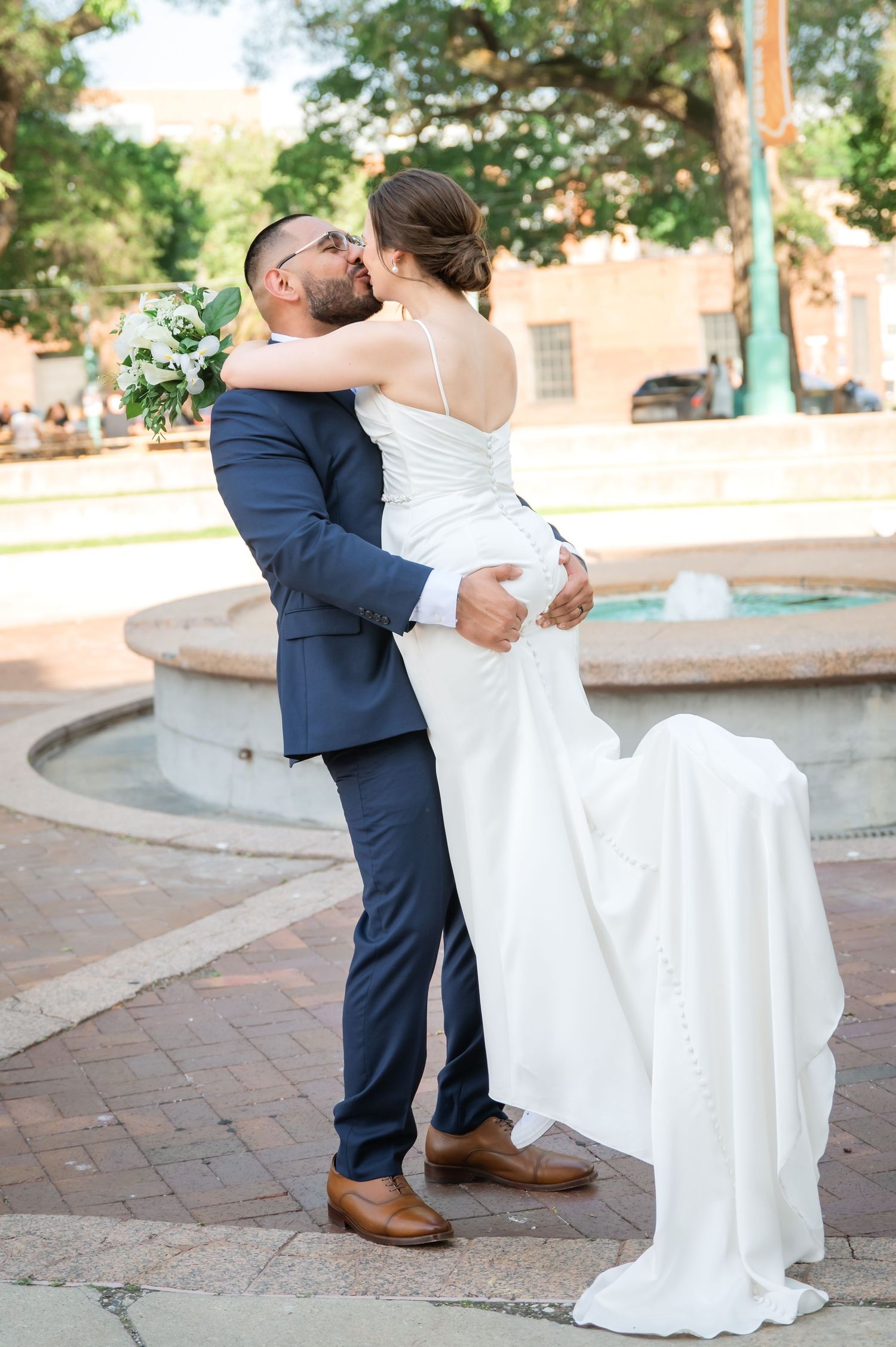 Bride in a lace wedding gown and groom in a gray suit embrace on a park path with a grassy lawn and city skyscrapers behind them.