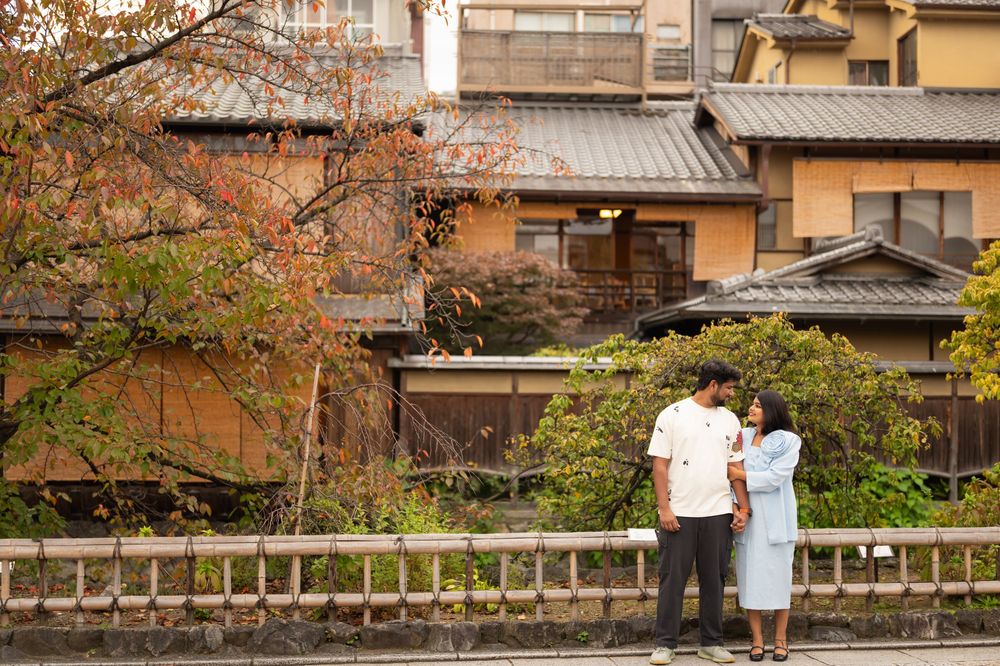 Couple stands together along a wooden railing in Kyoto's Gion district, with traditional Japanese wooden houses and autumn foliage behind.