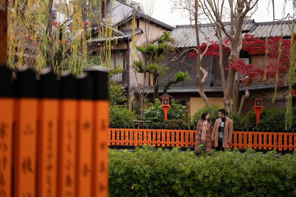 Two people stand near an orange wooden fence in front of traditional Japanese houses, red autumn foliage and stone lanterns in Kyoto.