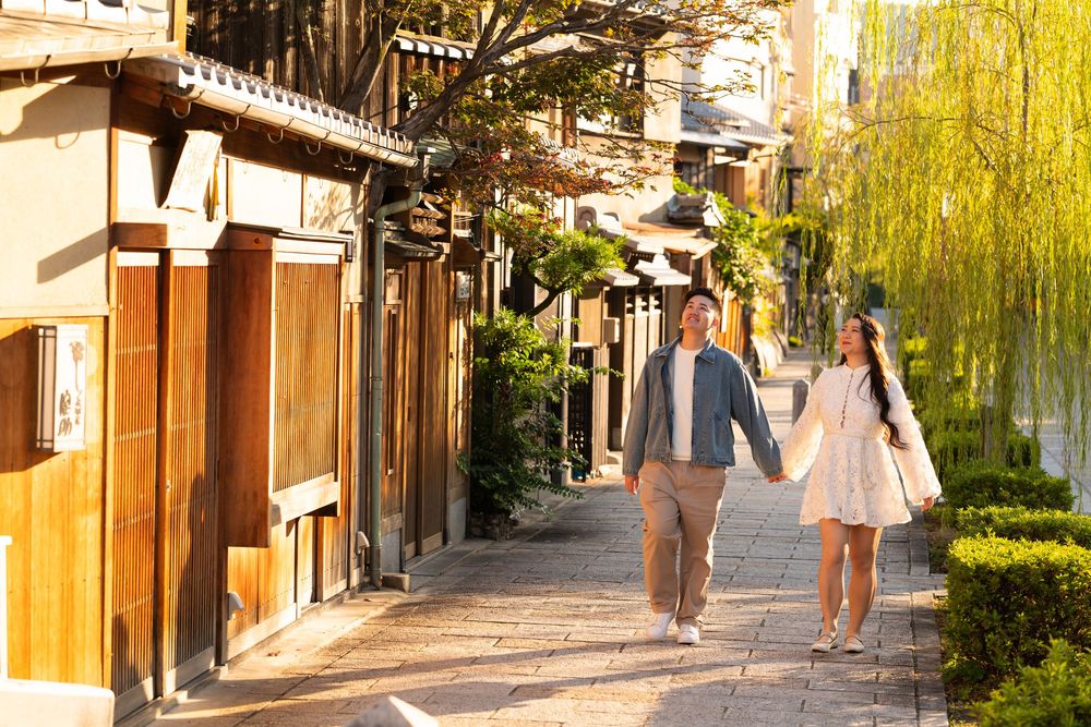 Couple holding hands strolling along a sunlit cobblestone path beside traditional wooden Japanese buildings and lush willow trees.