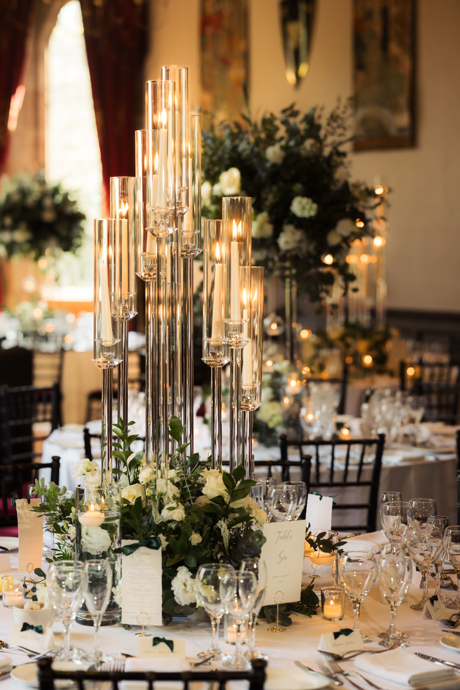 Elegant wedding reception table with tall glass candelabra, white floral centerpieces, greenery, and crystal glassware in a grand ballroom.