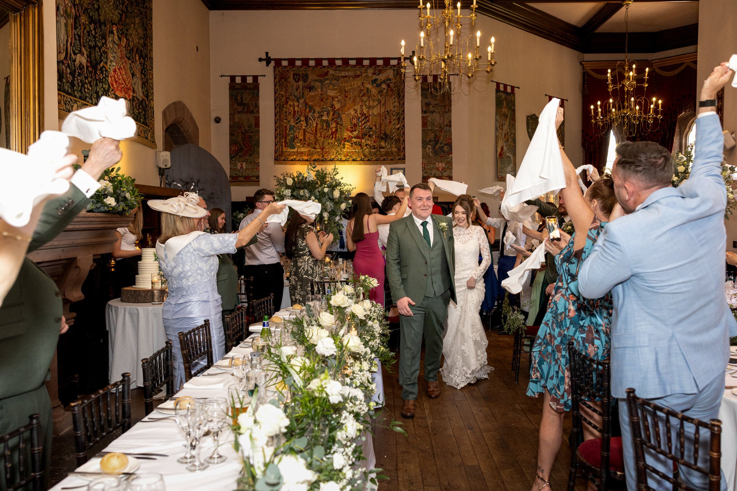 Bride and groom walk down the aisle of a rustic reception hall as wedding guests wave white napkins beside tables with floral centerpieces.