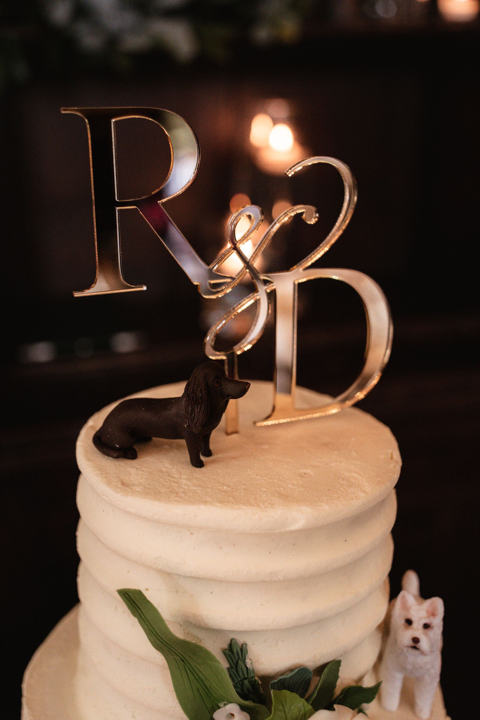 Wedding cake with textured white buttercream, a mirrored gold R and D monogram topper, and dachshund and white terrier dog figurines.