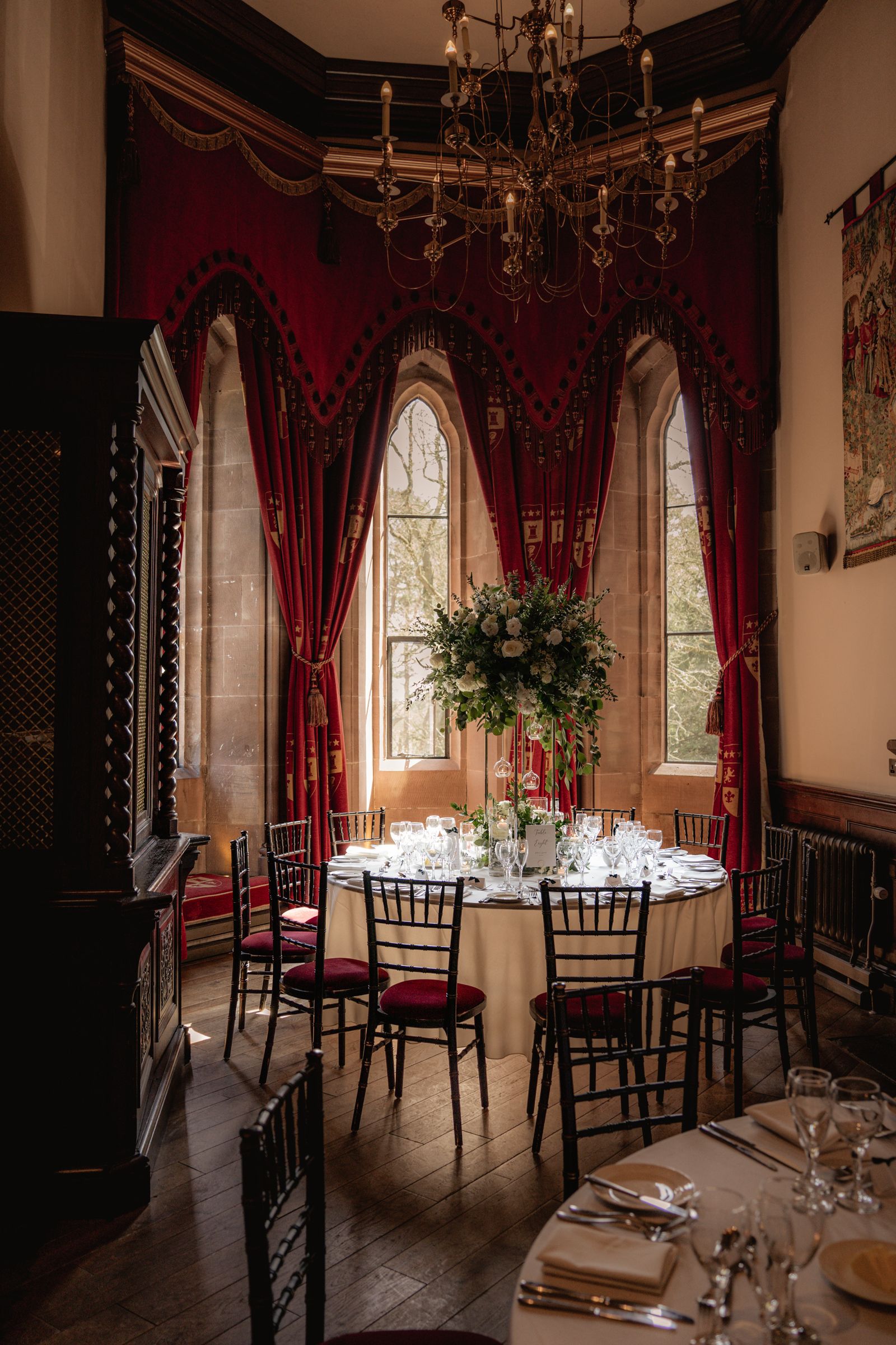 Elegant round banquet table with floral centerpiece in a castle room with red velvet drapes, arched windows, and a chandelier.