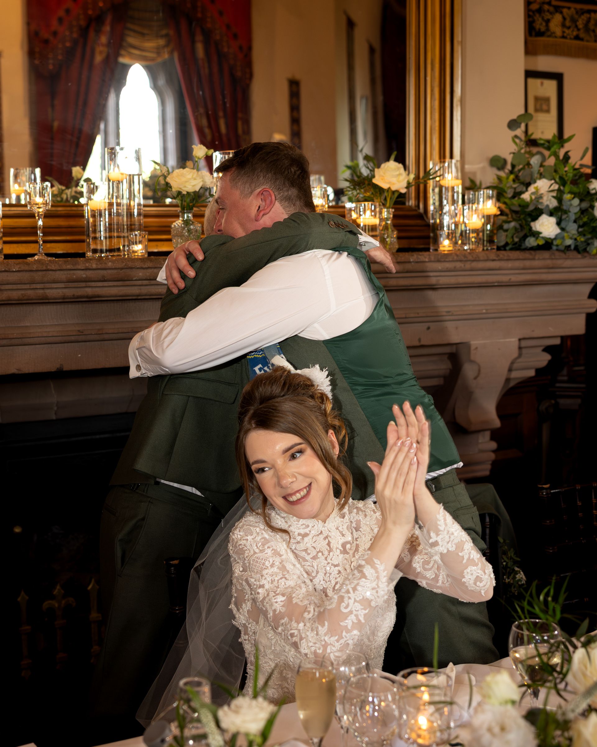 A smiling bride in a lace wedding dress applauds while a groom in a green vest hugs a man beside a candlelit table with flowers.