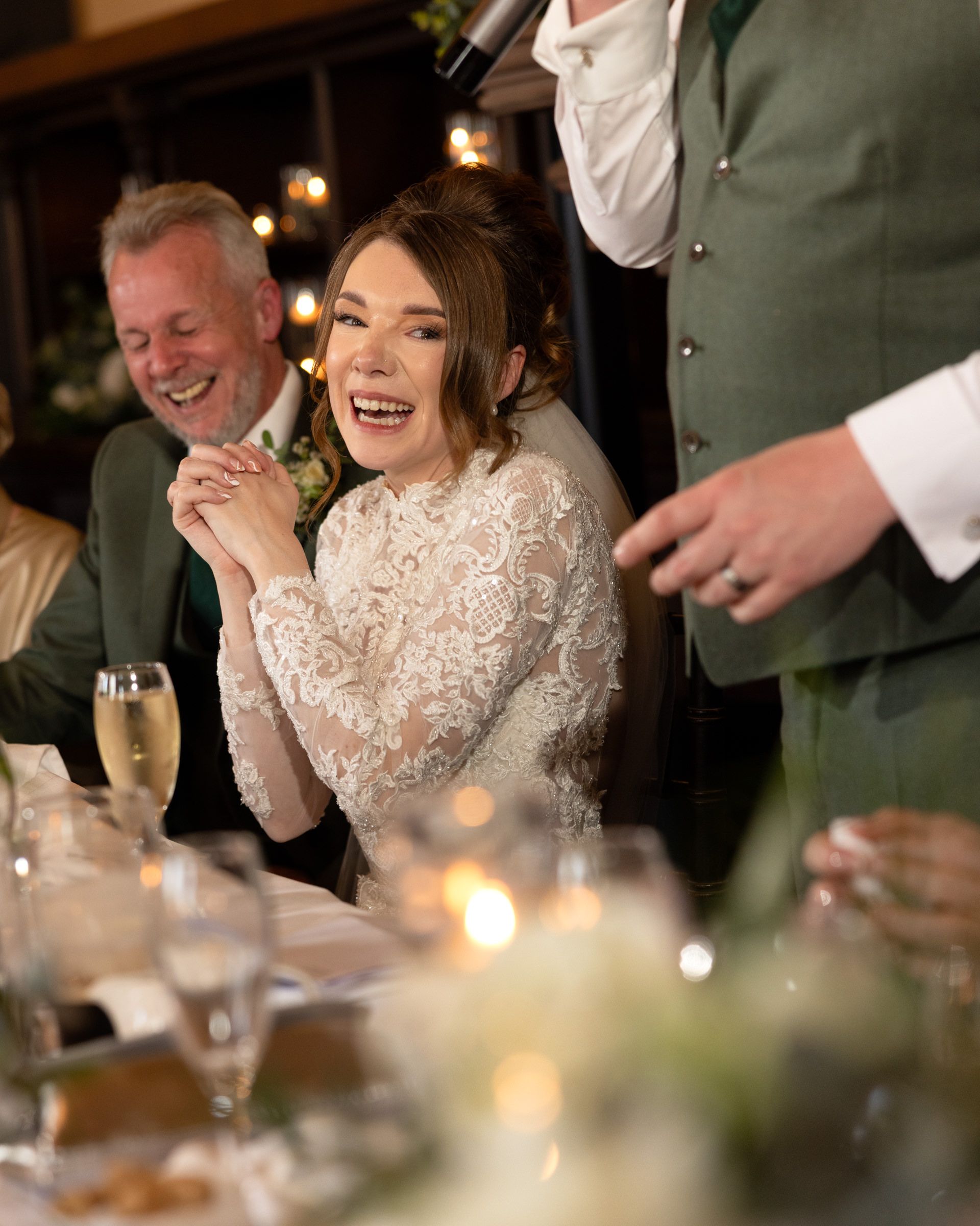 Smiling bride in a lace wedding dress laughs at the reception table during a speech, with candles and champagne glasses nearby.