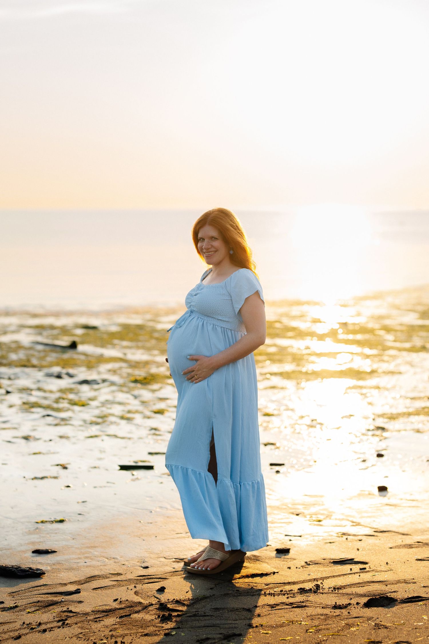 Pregnant woman in a flowing light blue dress cradles her belly while standing on a wet sandy beach at golden sunset.