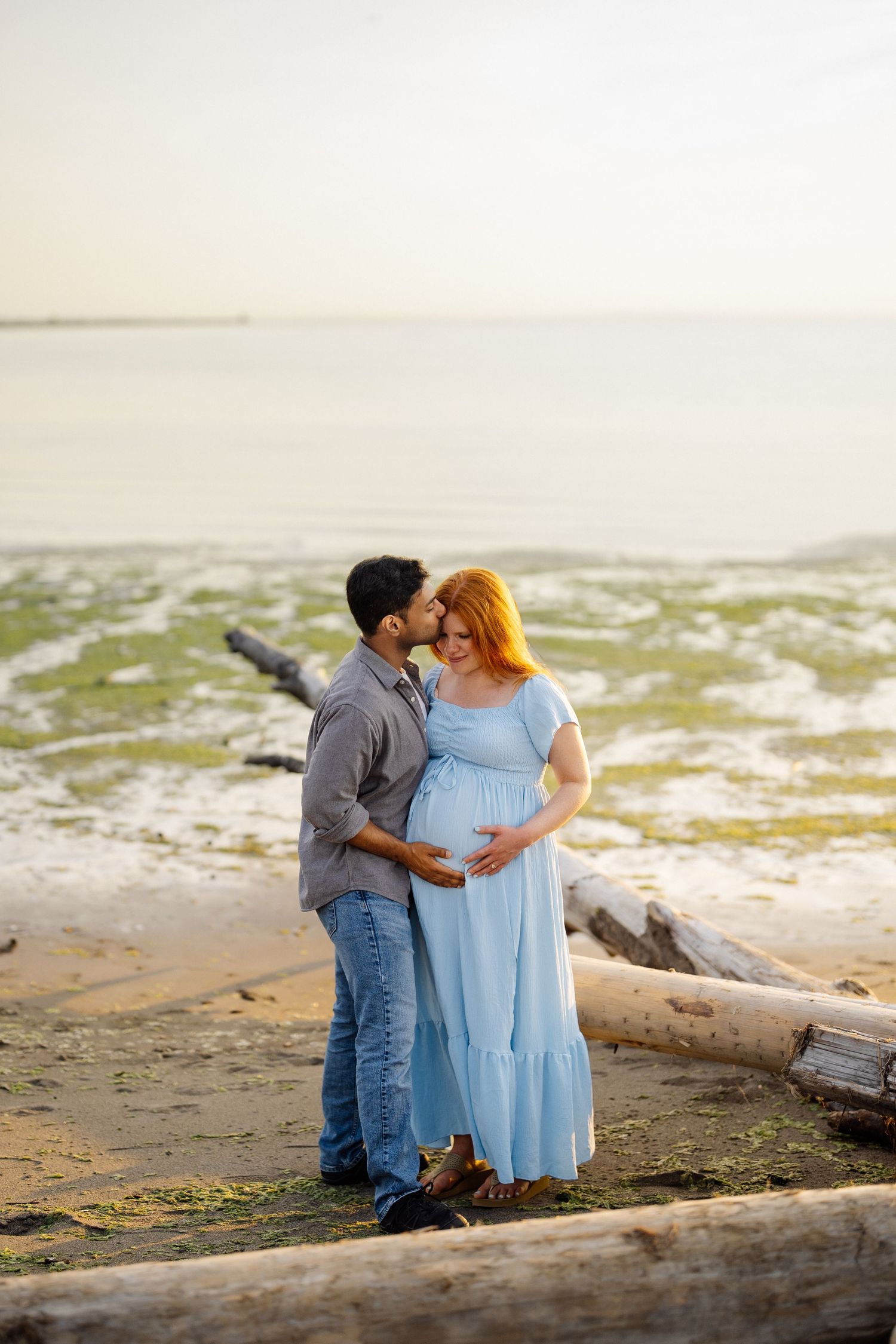 Expecting couple shares a tender kiss on a sandy beach with driftwood, her light blue dress hugging her baby bump at sunset.
