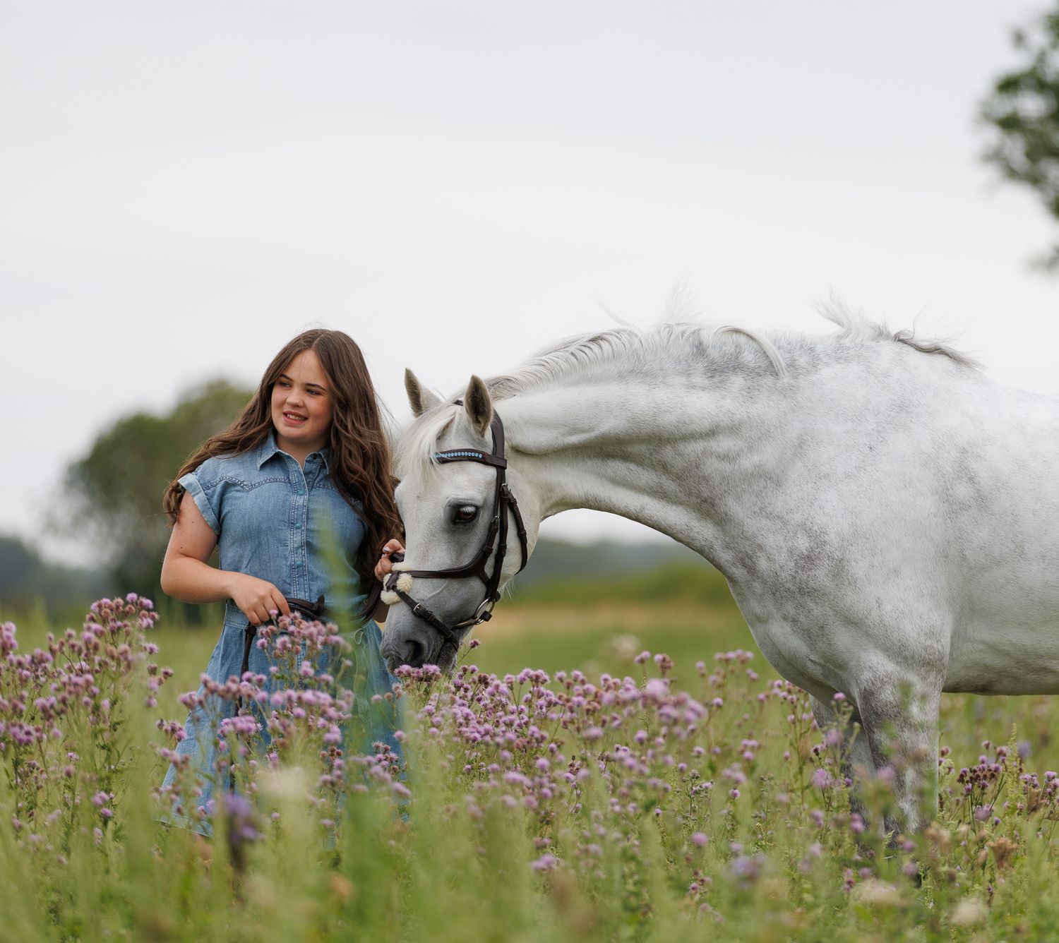 A woman sits on a log beside a brown horse in a field, holding its reins in a serene black and white photo.