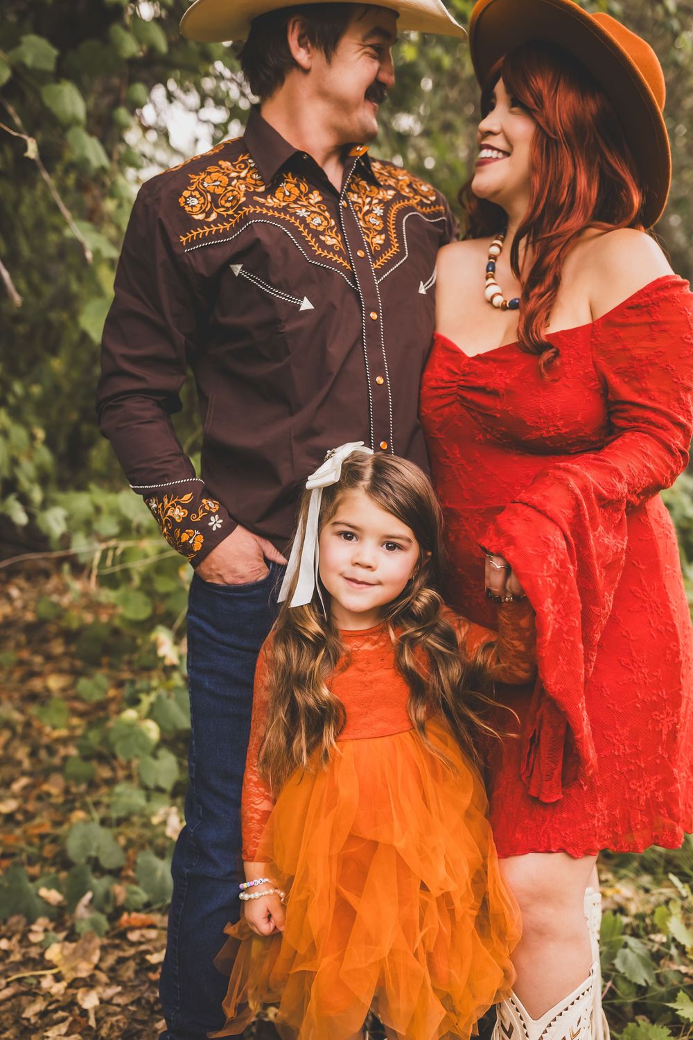 Parents in western wear smile at their daughter in an orange tutu dress outdoors.