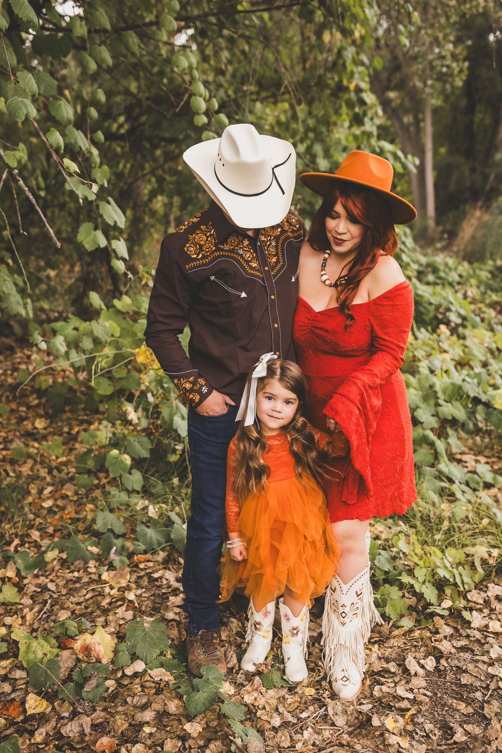 Family of three in Western attire poses outdoors in fall foliage, wearing cowboy hats and boots.