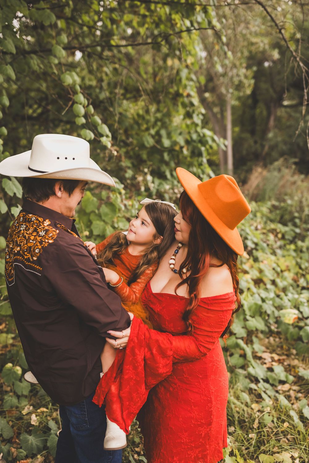 Family of three in western attire, parents in cowboy hats holding young girl outdoors.