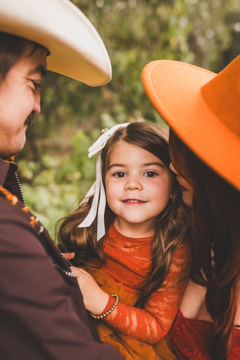 Smiling young girl with white bow held by parents wearing cowboy hats outdoors.
