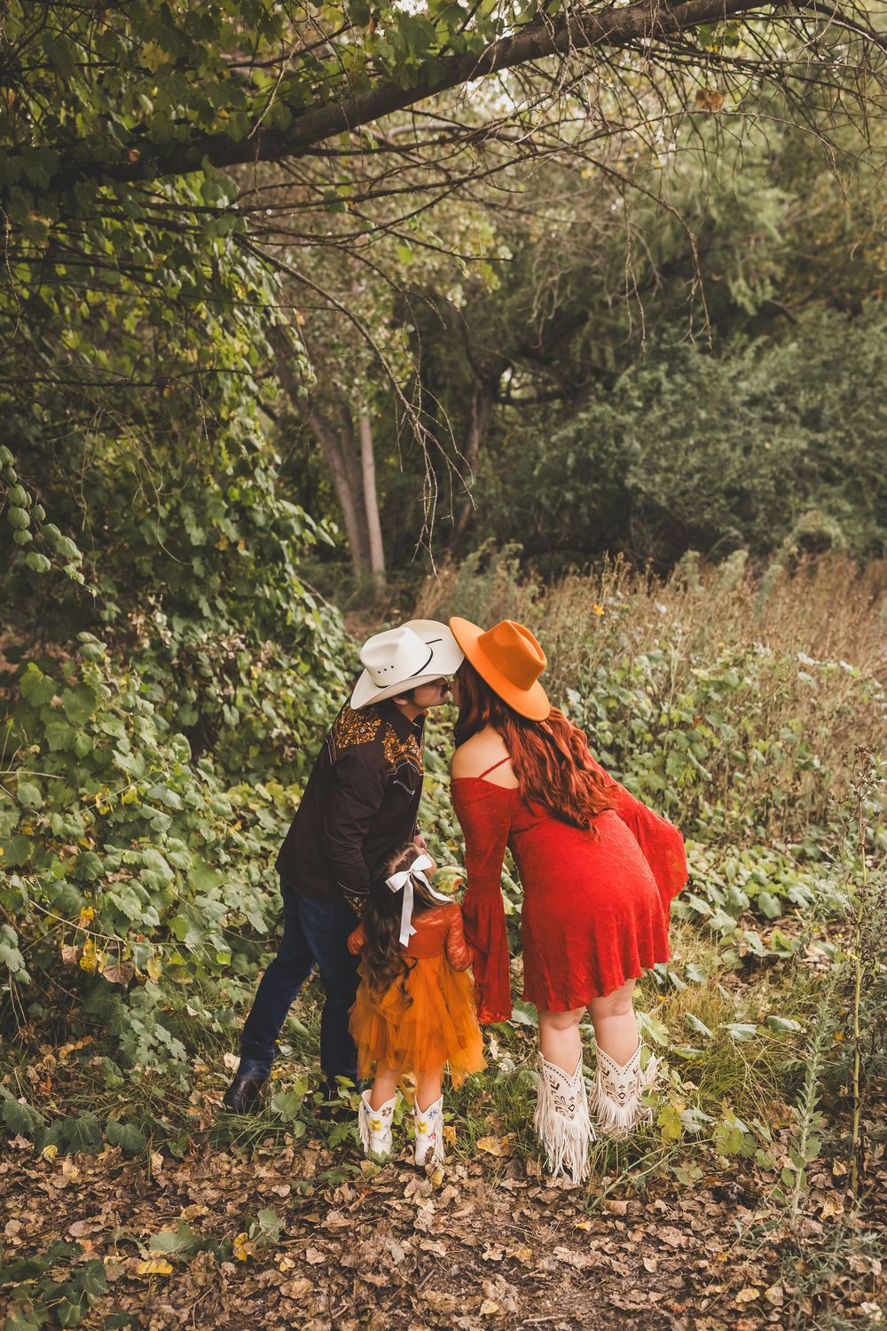 Two women in cowboy hats and boots share a kiss outdoors, one in red dress and one in black.